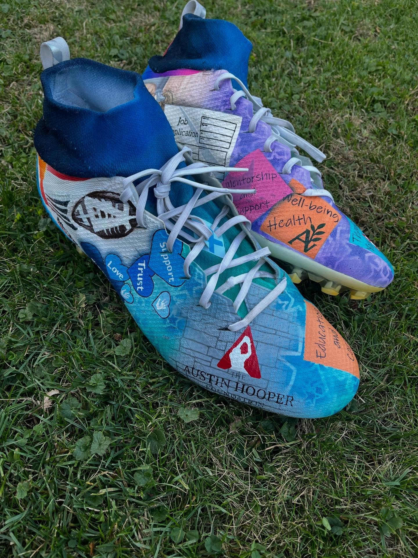 Colorfully decorated athletic shoes with text and symbols promoting health, mentorship, and support, lying on grass.