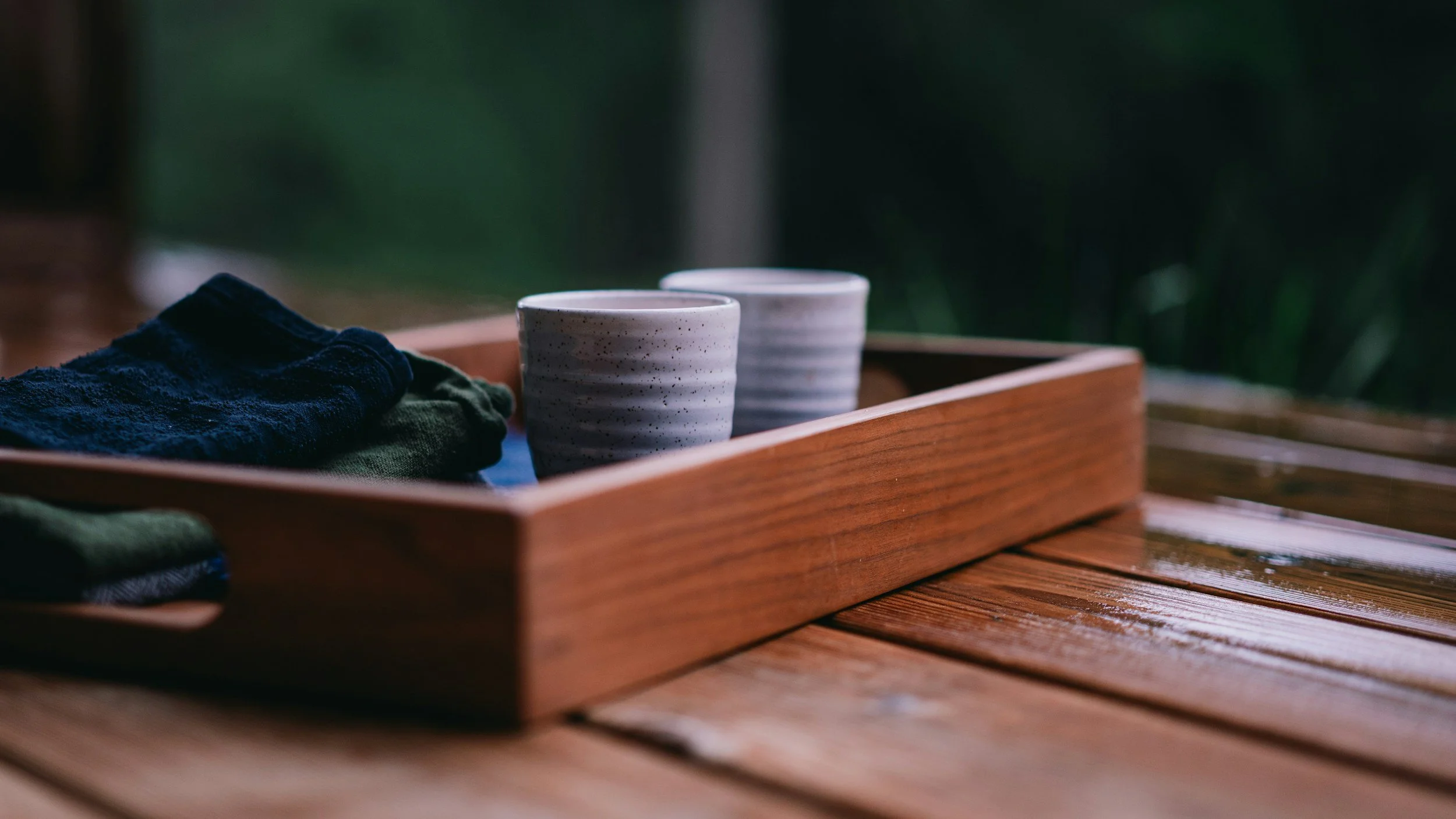 A wooden tray with two ceramic cups and some folded clothes on a wet wooden surface outdoors.