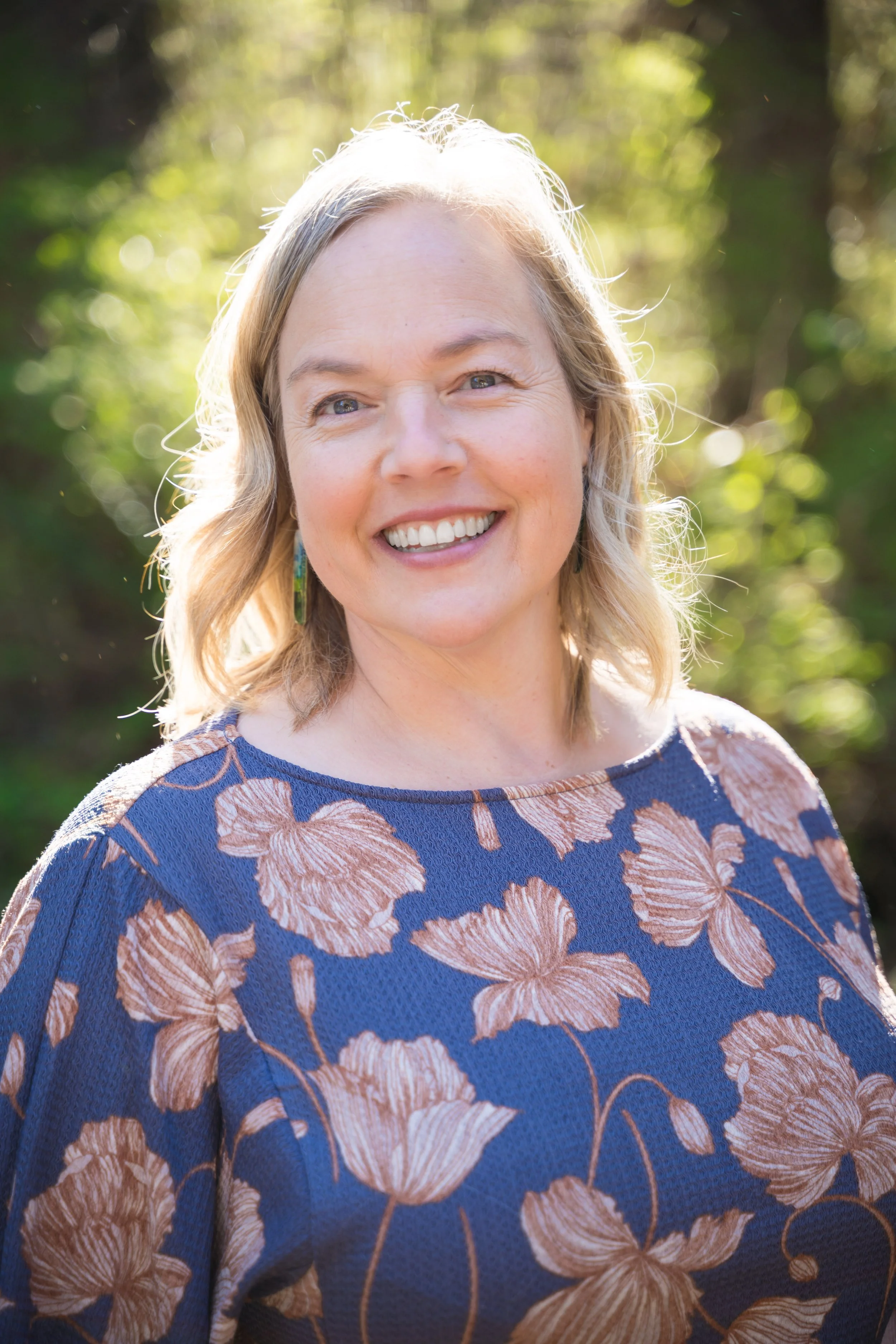 A smiling woman with blonde hair wearing a blue floral top outdoors with sunlight and blurred green trees in the background.