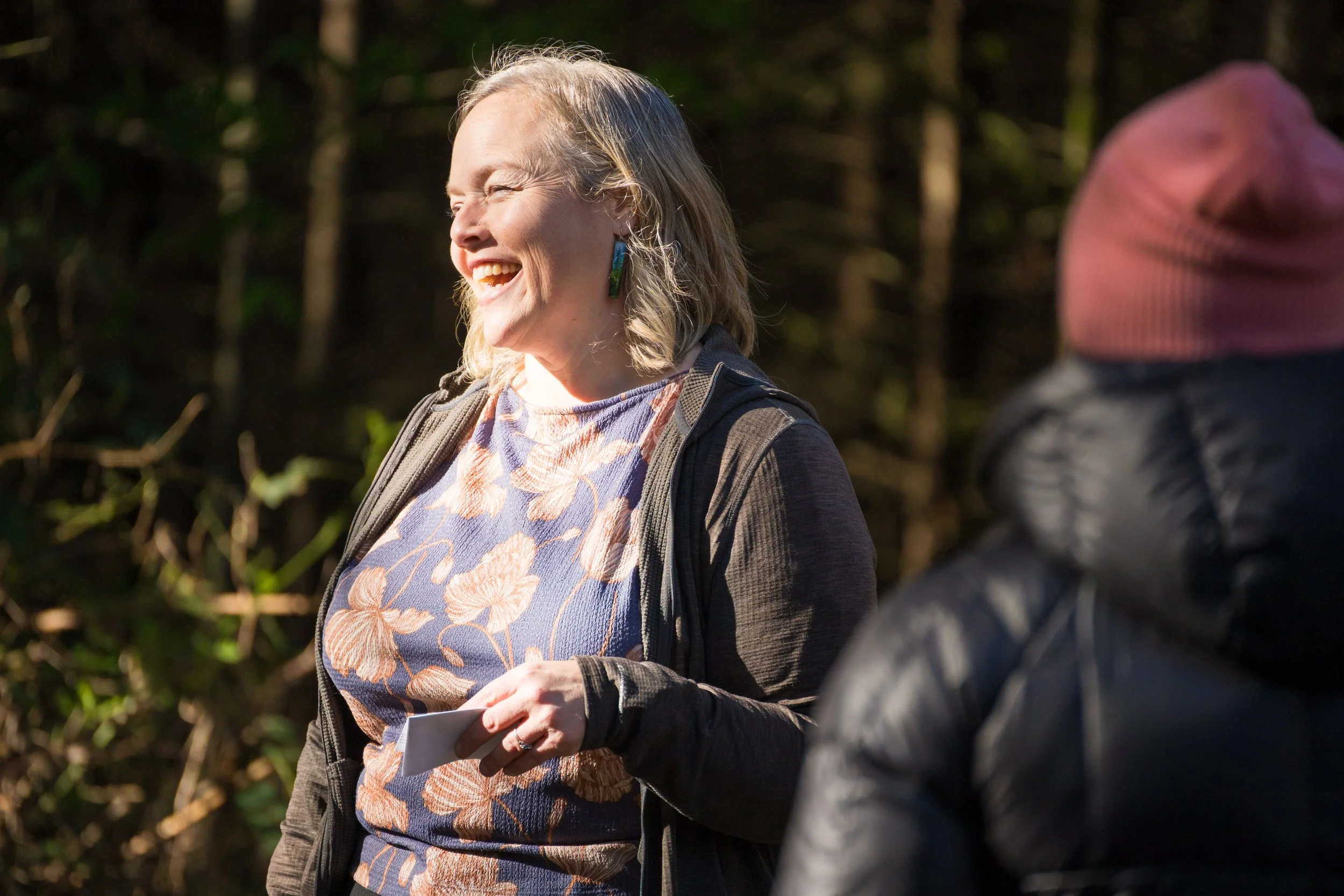 A woman laughing outdoors with sunlight on her face, wearing a patterned shirt and dark jacket, holding a paper, standing in a forested area.