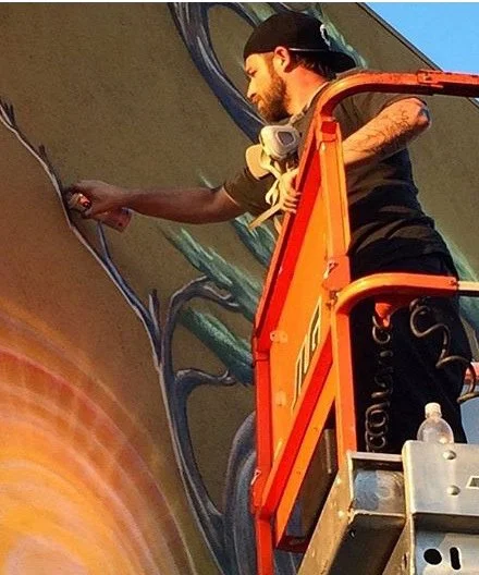 An artist is painting a large mural on a wall from a scissor lift. The artist has a beard, tattoos, and is wearing a cap and a headset around his neck.