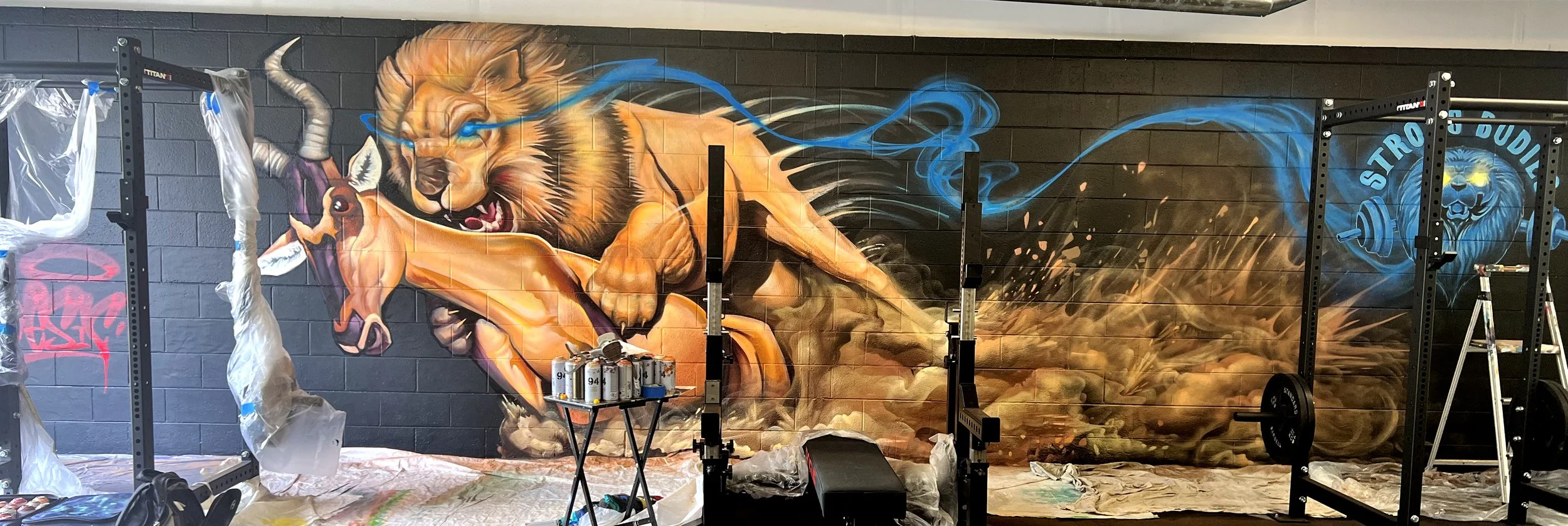 A large mural of a fierce lion with glowing blue eyes and flowing blue smoke on a black brick wall, with gym equipment and ladders in the foreground.