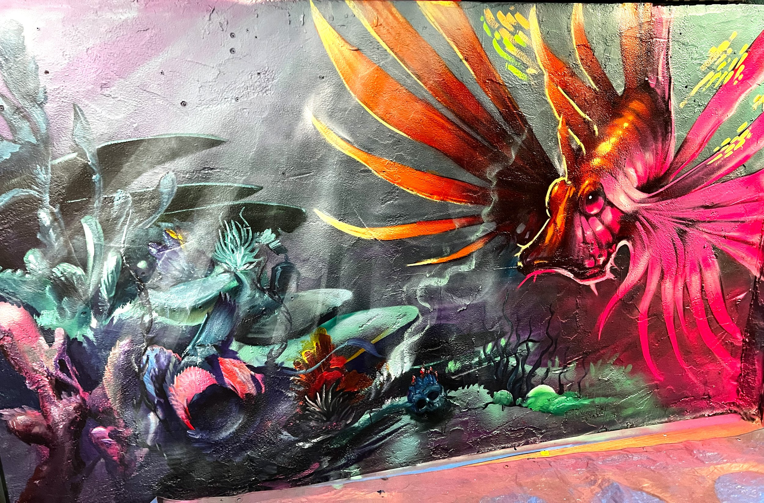 Vibrant graffiti mural featuring a large pink and orange insect or alien creature with elongated limbs on the right side, and abstract plant-like shapes in shades of green, blue, and pink on the left side.