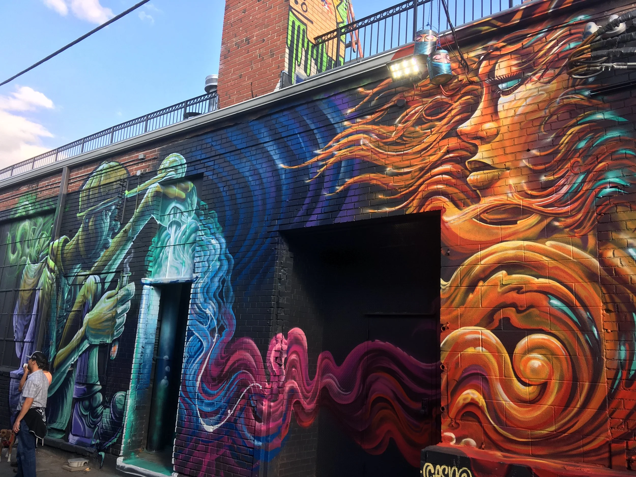 Colorful mural of a woman with flowing hair on a brick building wall, with a doorway incorporated into the artwork, and people standing nearby.