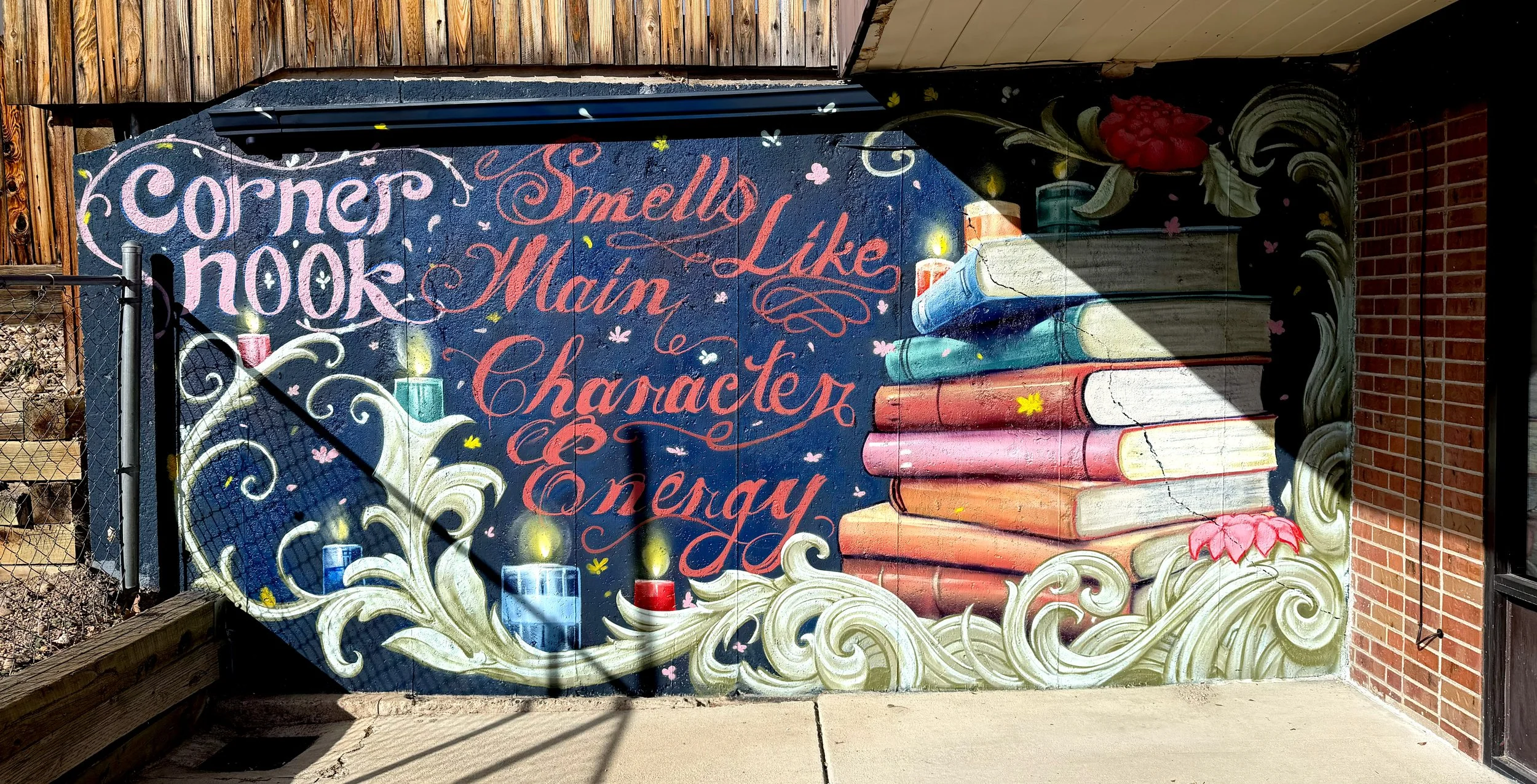 Colorful mural with books and candles surrounding the slogan 'Corners Nook Smells Like Main Character Energy' painted on an outdoor wall.