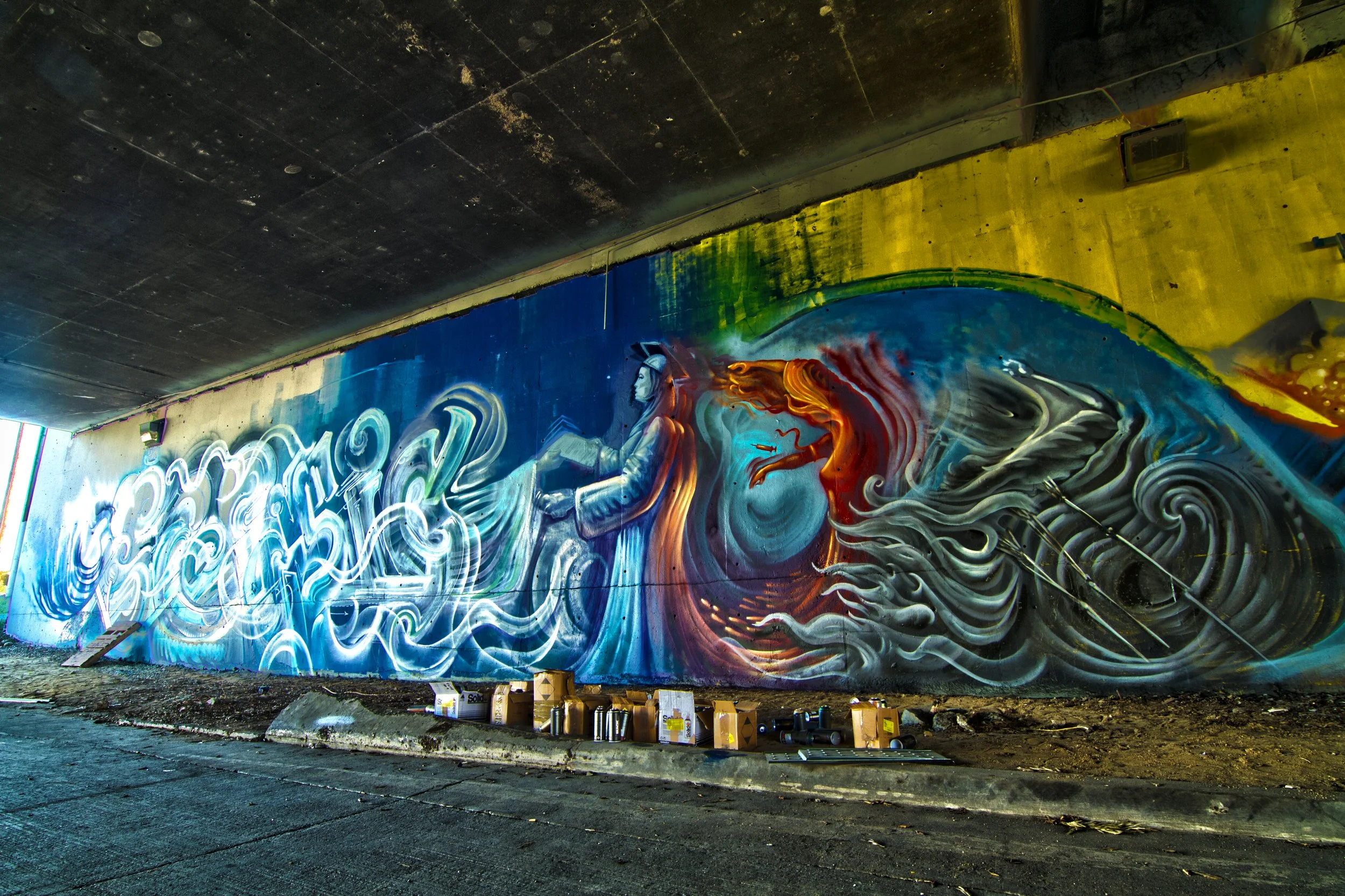 Colorful mural graffiti on a large wall beneath a bridge, depicting a woman with long hair holding an open book, surrounded by abstract swirling shapes and patterns.