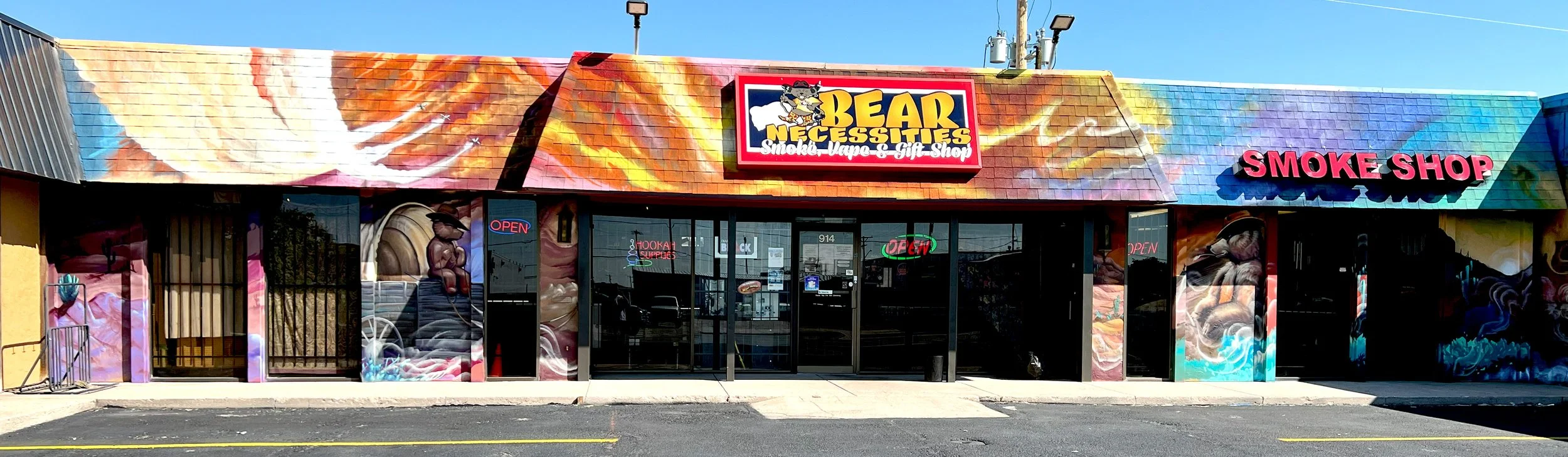 Front of a store with colorful murals, signage for Bear Necessities Smoke, Vape & Gift Shop, and a separate sign for Smoke Shop. The storefront has glass doors with illuminated 'Open' signs and an 'Open' neon sign outside.
