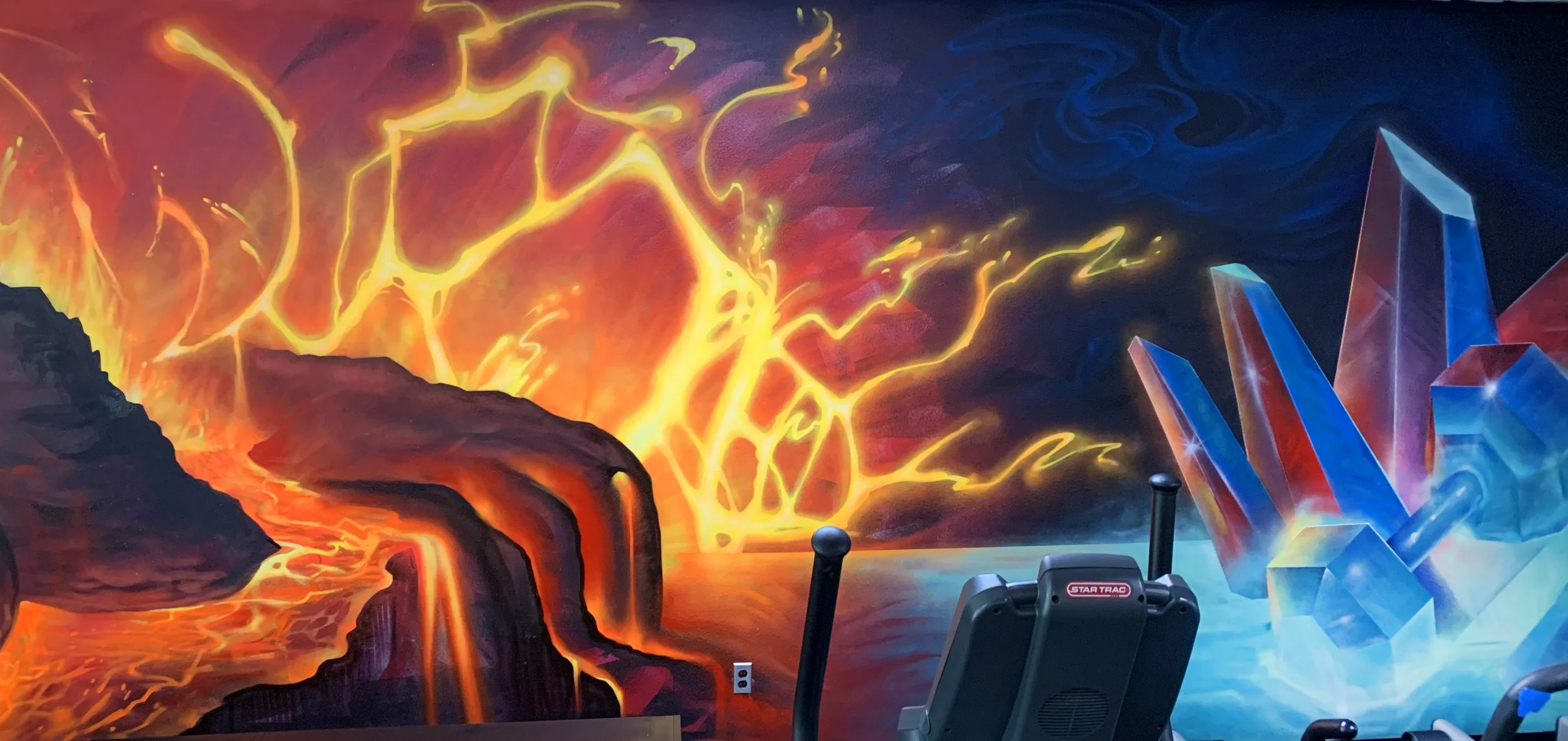 Colorful wall mural depicting a volcanic eruption with flowing lava on the left, and a cluster of glowing blue and red crystals on the right. There are gym equipment and electrical outlets in front of the mural.
