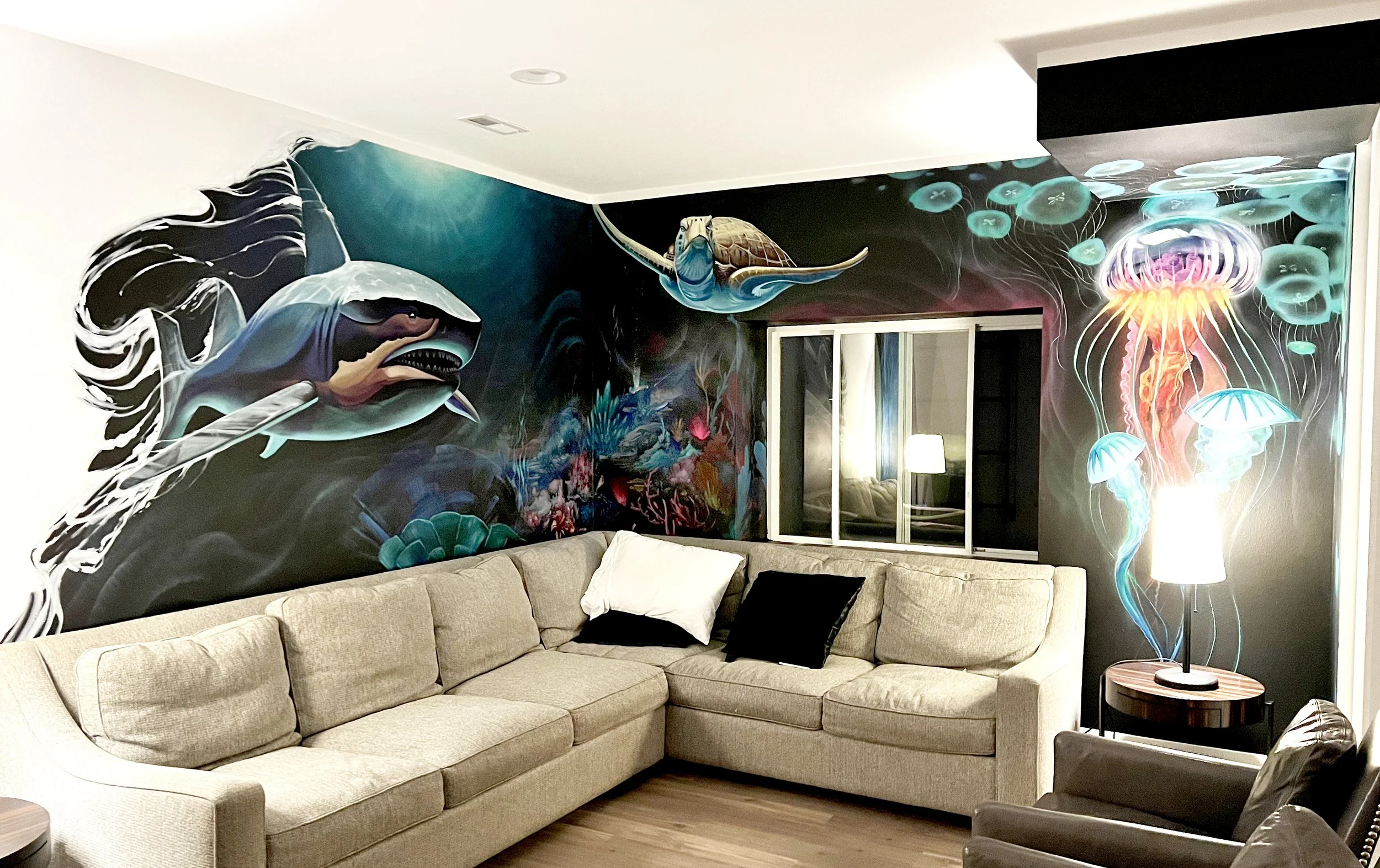 Living room with a beige sectional sofa, black and white throw pillows, and an underwater-themed mural featuring a shark, a turtle, and jellyfish on the wall.