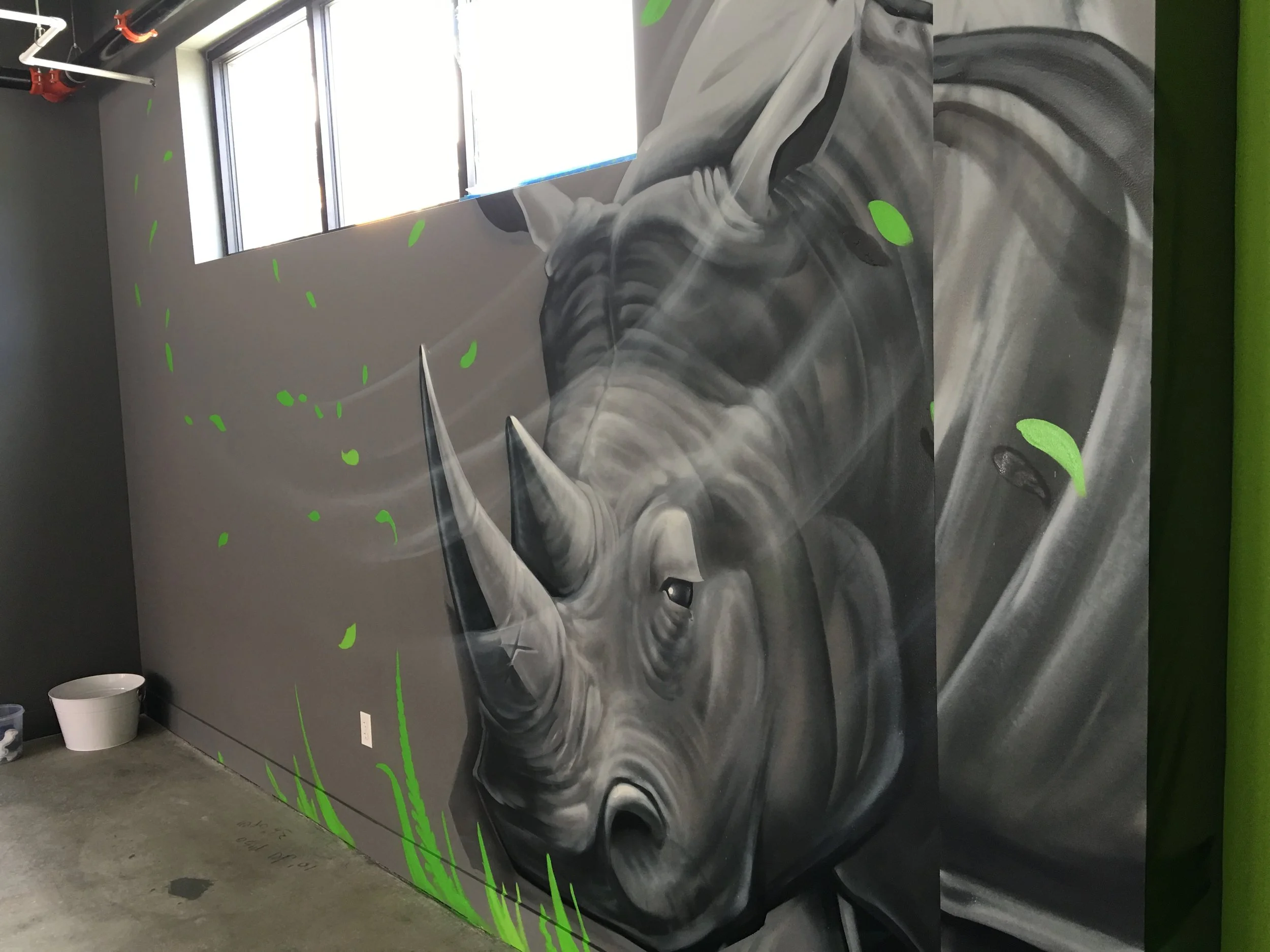 A detailed mural of a rhinoceros painted in shades of gray with neon green accents on a wall in an indoor space with a window.