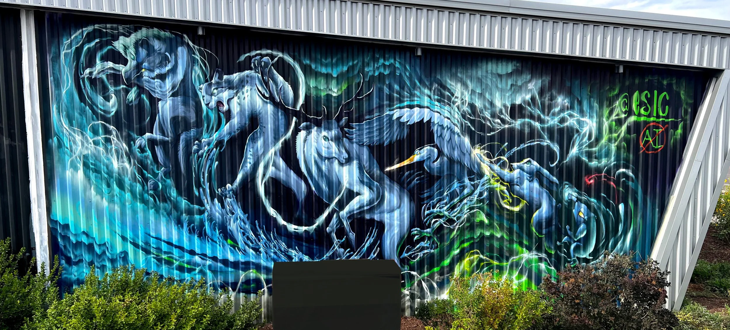Graffiti mural of mythical animals including a lion, eagle, and deer on a metal wall with trees and bushes in front.
