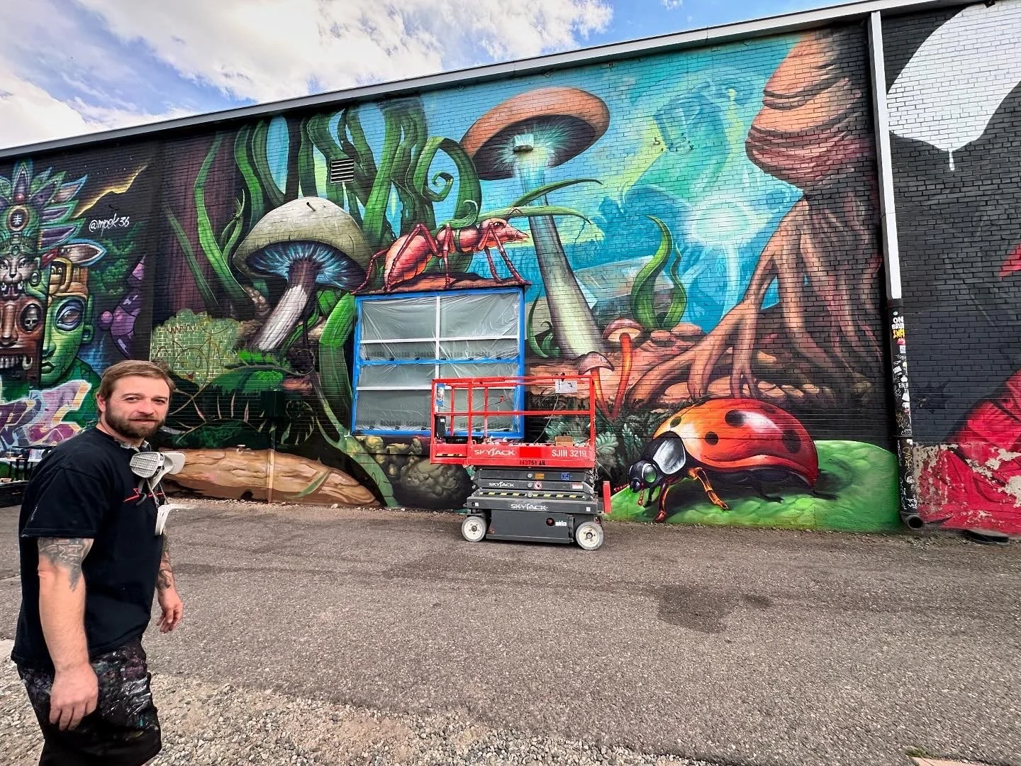 Street artist standing in front of a large colorful mural on a brick wall, depicting fungi, a ladybug, and various abstract and fantasy elements. Art supplies and a lift platform are placed in front of the mural.