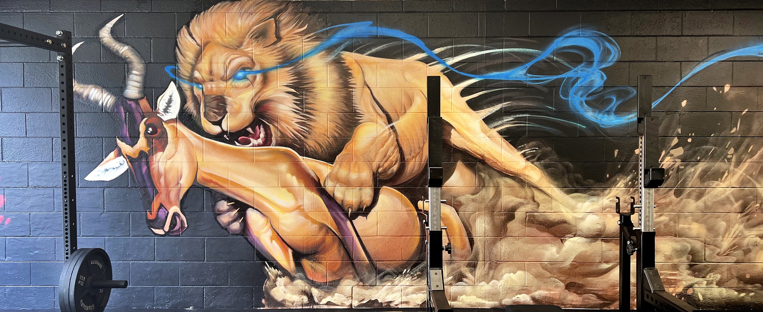 Colorful mural of a lion and a goat with blue smoke and dynamic background on a black brick wall, in a gym setting with workout equipment.
