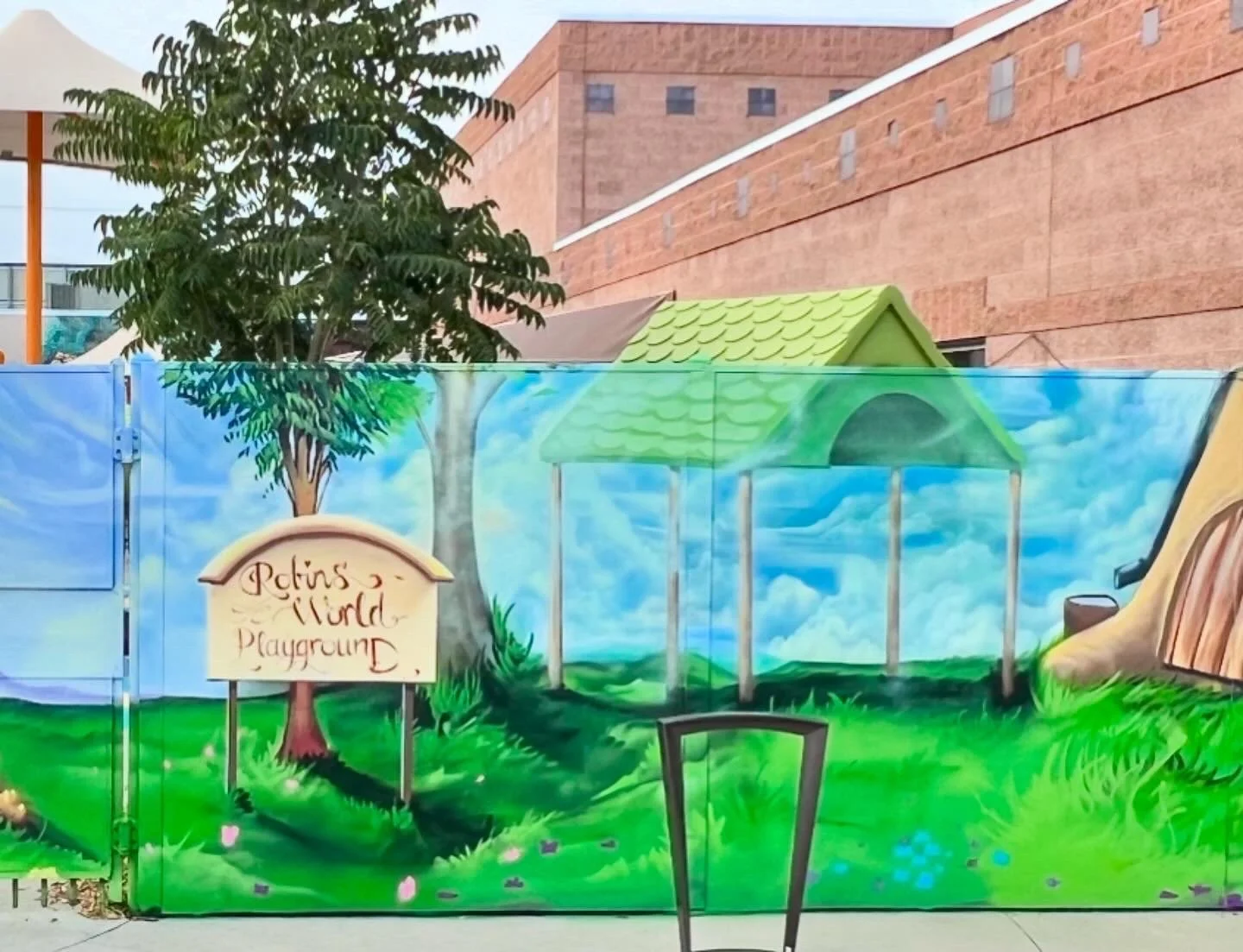Colorful mural of a playground on a fence, including a tree, a sign that reads 'Petins World Playground', a green-roofed play structure, a slide, and grass and clouds painted in the background.