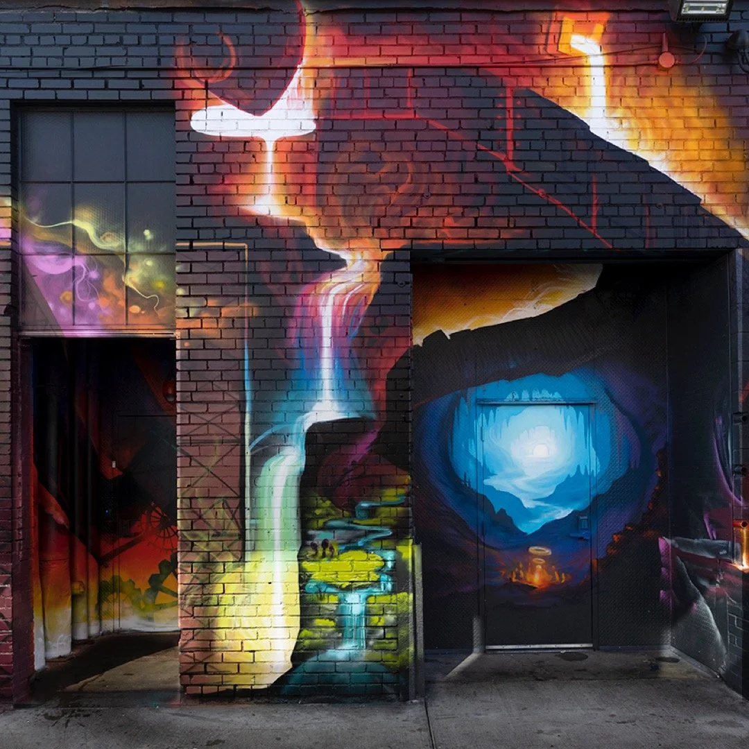 Colorful mural on a brick wall depicting a face with a waterfall flowing from the eye into a stylized landscape of mountains and a cave with a blue circular opening and a bright light shining through.