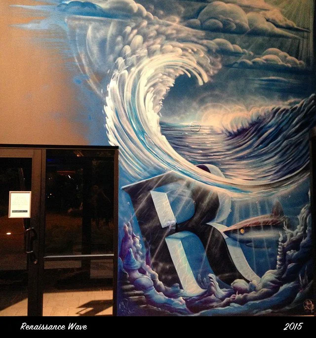 A large mural depicting a massive ocean wave with clouds in the sky above, painted in shades of blue, white, and gray. The mural includes the letters 'R' and 'A' in the foreground, and there is a shark fin emerging from the water on the right side. T