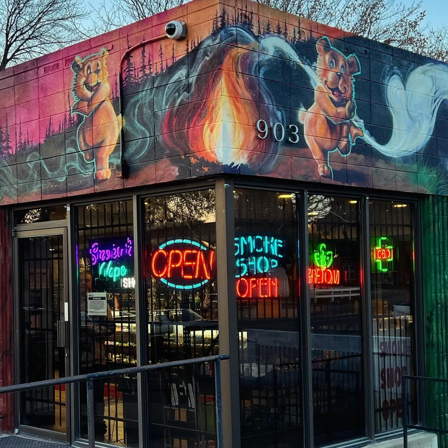Storefront with colorful mural of bears, fire, and nature on the upper exterior wall, and neon signs inside the store displaying 'Open,' 'Smoke Shop,' and other words. Glass doors and windows show the interior of the shop.