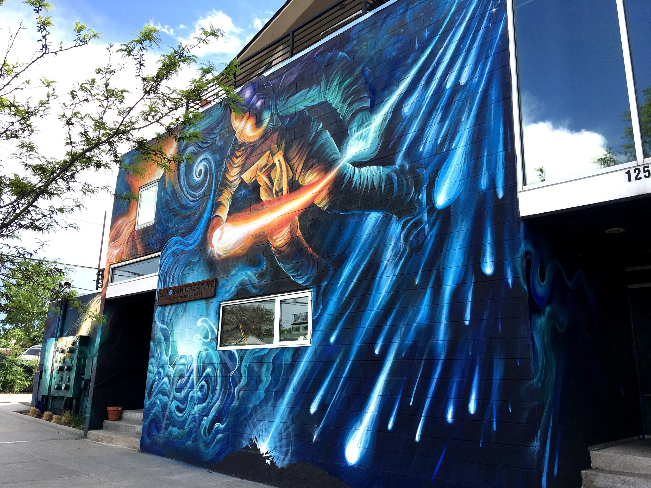 Colorful mural of a figure with a fiery sword, surrounded by swirling blue and orange abstract patterns on the exterior wall of a building.