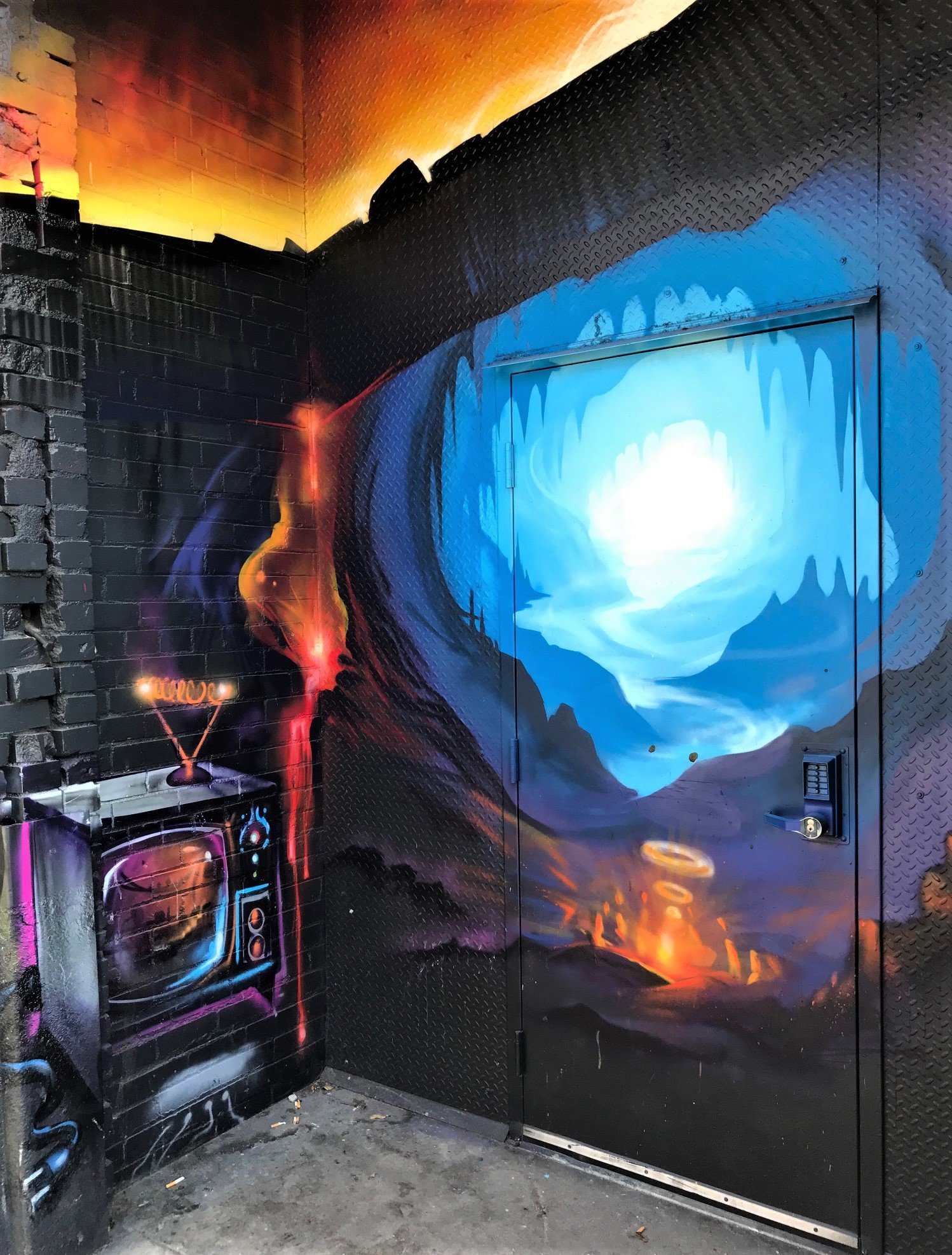 Colorful street art mural depicting a lava lamp, an explosion of blue light resembling a volcanic eruption, with a mountainous landscape and swirling clouds, painted on a black textured wall.
