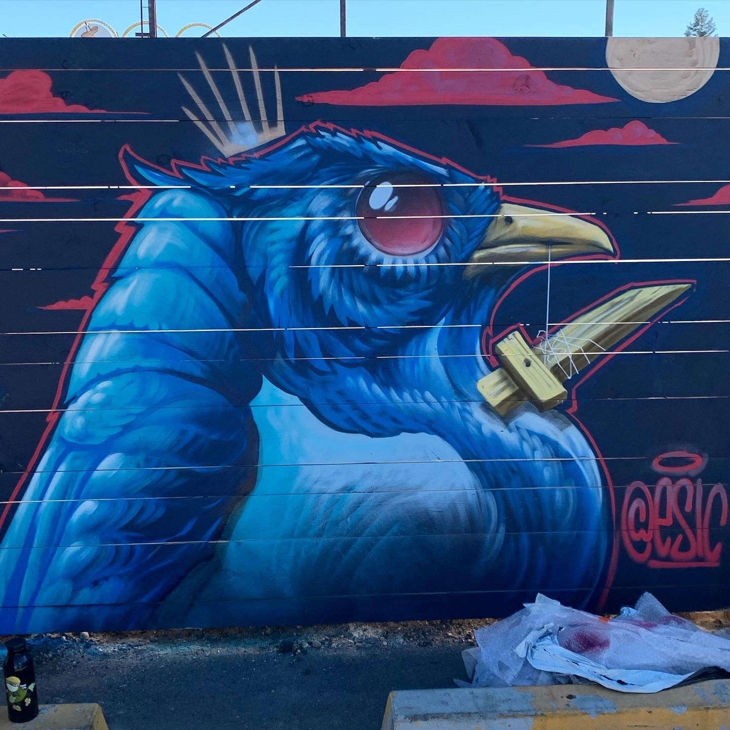 A graffiti mural of a blue bird with red and blue feathers, holding a yellow sword in its beak, with pink clouds and a moon in the background.