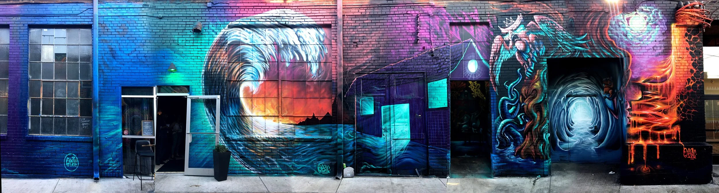 Colorful mural on a brick wall featuring a large wave, sunset, geometric patterns, a dragon, and cosmic elements.