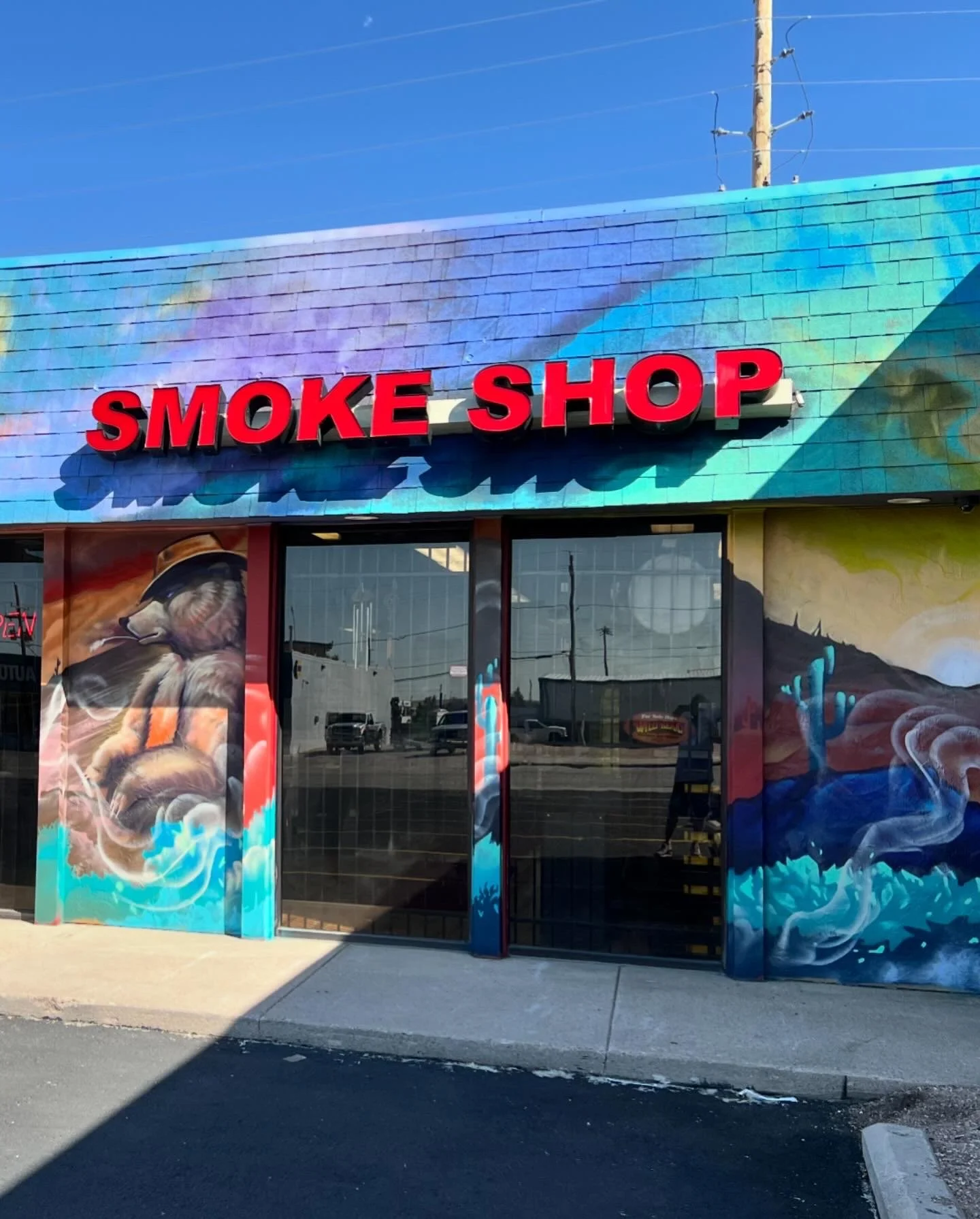 Exterior of a retail store with a blue-tiled roof, red sign reading 'SMOKE SHOP', and colorful artwork of a dog and smoke on the walls.