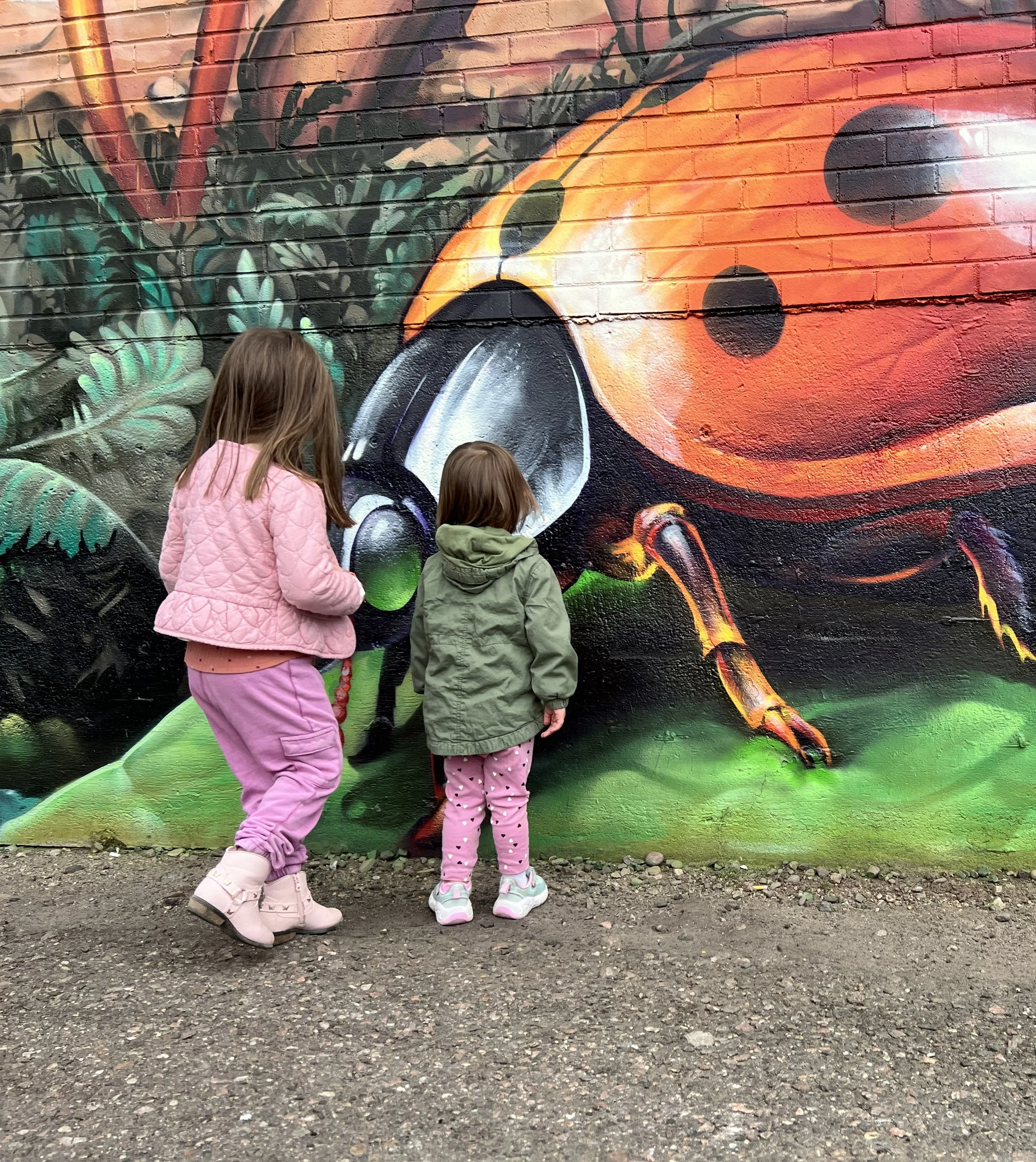 Two young girls looking at a colorful mural of a ladybug on a brick wall.