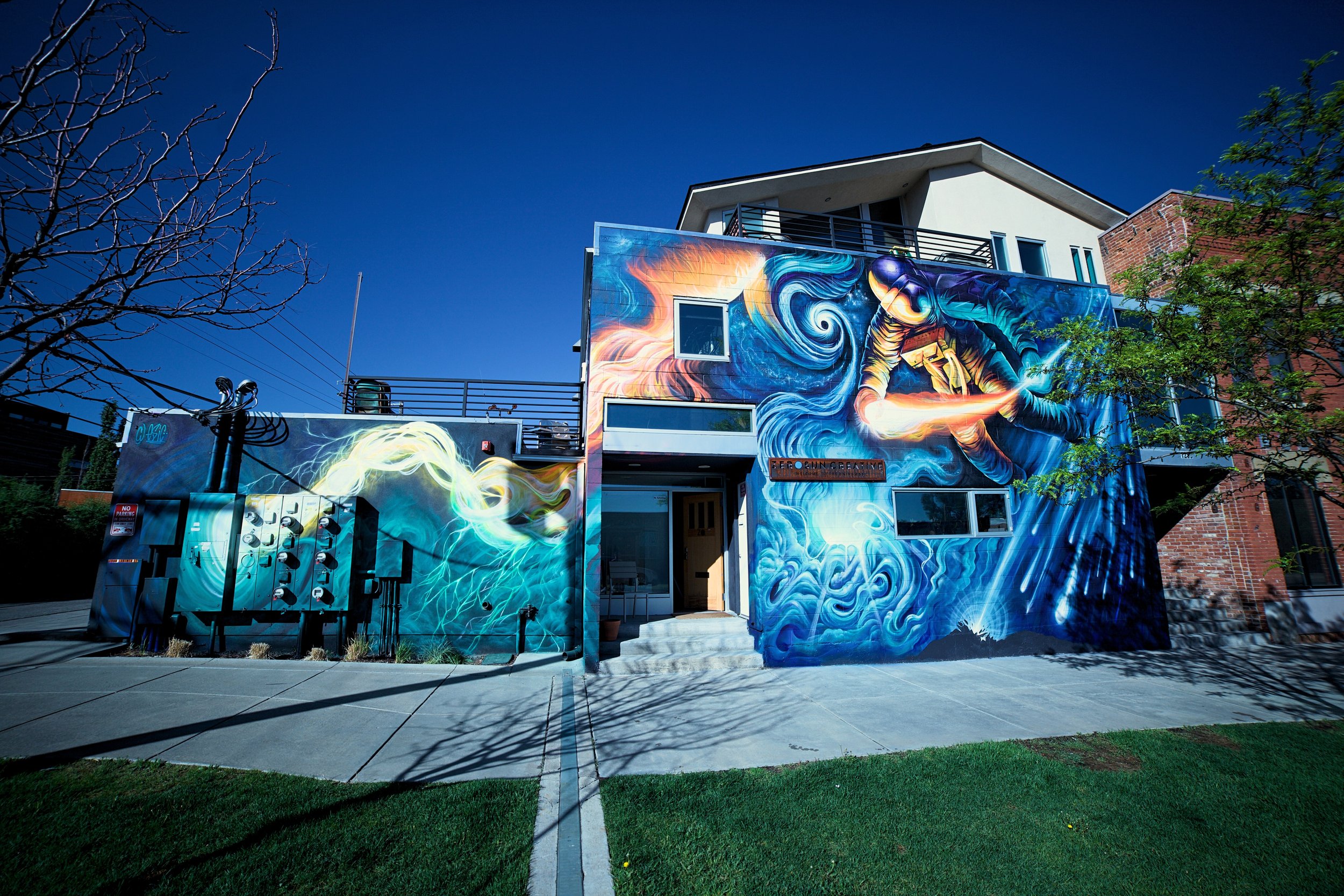 Colorful mural on the exterior of a building, depicting a figure in space helmet with swirling blue and orange effects, and a wall with electrical equipment.