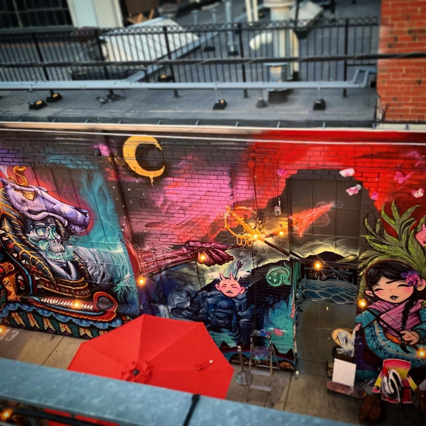 Colorful graffiti art on a brick wall featuring characters and designs, including a skeleton with a snake, a pink cartoonish creature, and a girl with long dark hair and a pink dress, with lights and an umbrella in the foreground.