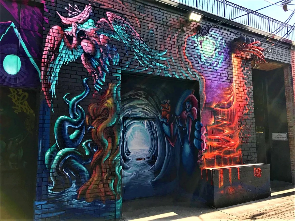 Colorful graffiti mural on a brick wall depicting a mermaid with long flowing hair, a swirling vortex, a tattooed woman, and vibrant abstract elements, with bright colors and detailed artwork.