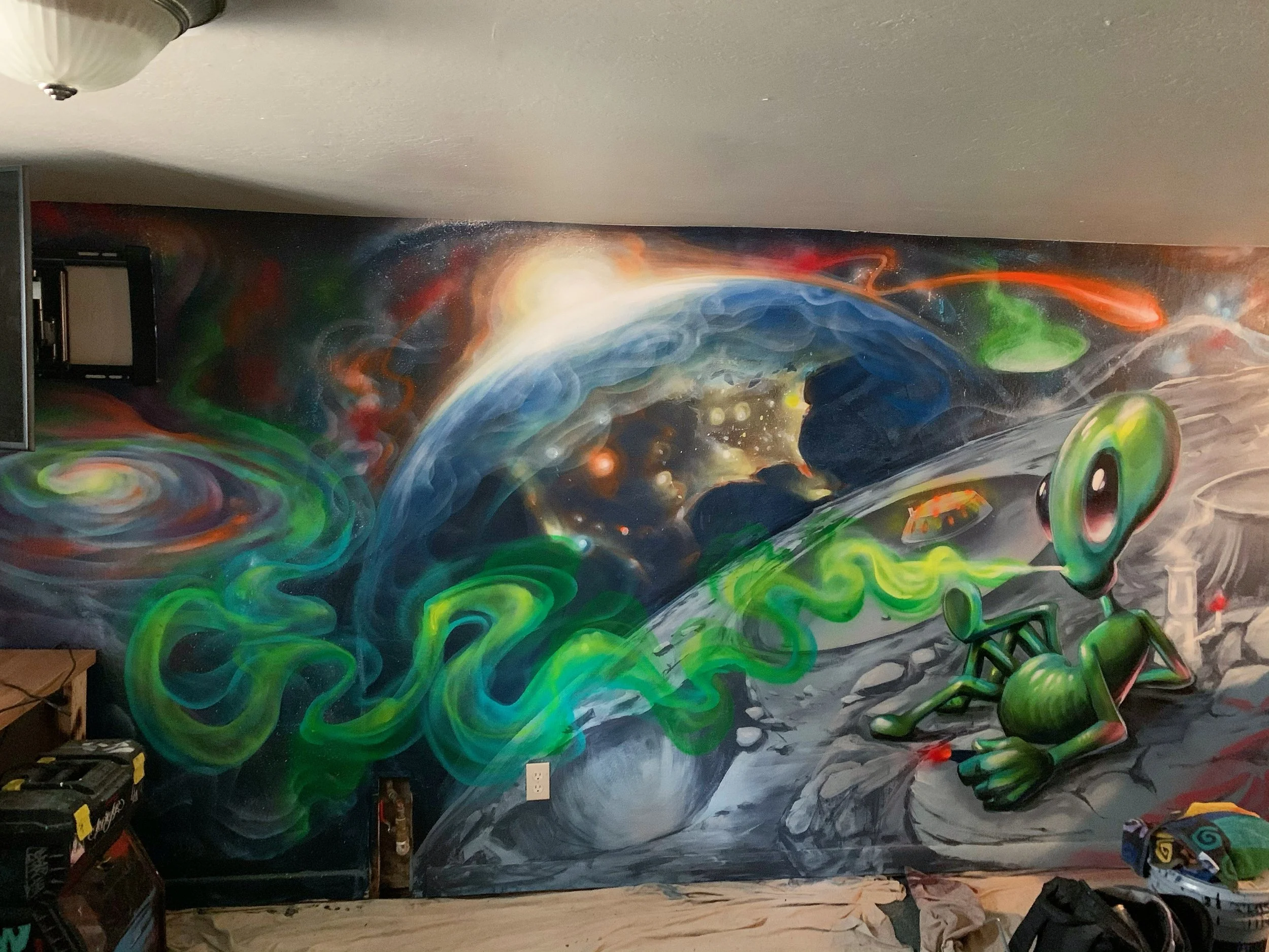 A space-themed graffiti mural with an alien character sitting on the moon, Earth in the background, swirling green and blue cosmic elements, and colorful stars and nebulae.