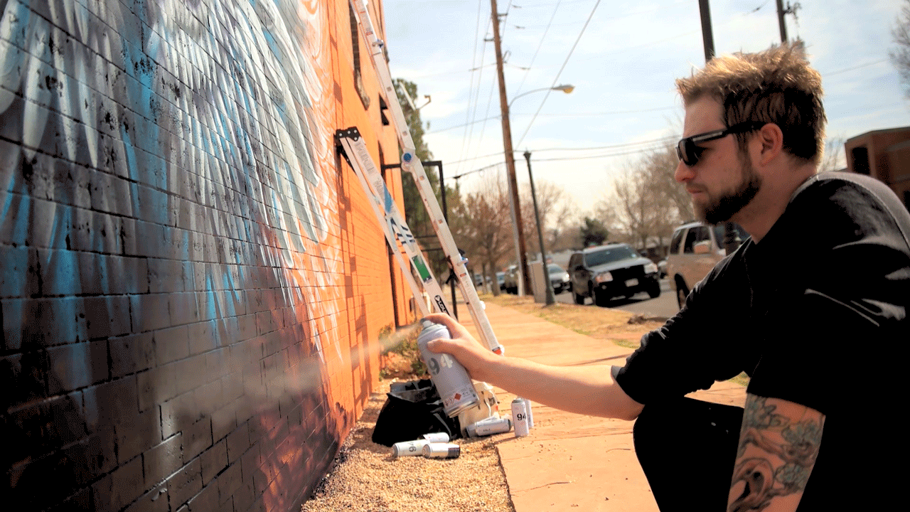 A man wearing sunglasses sprays graffiti on a brick wall.
