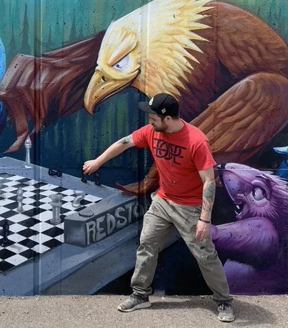 A man in a red t-shirt and beige pants standing in front of a mural with an eagle and animals playing chess
