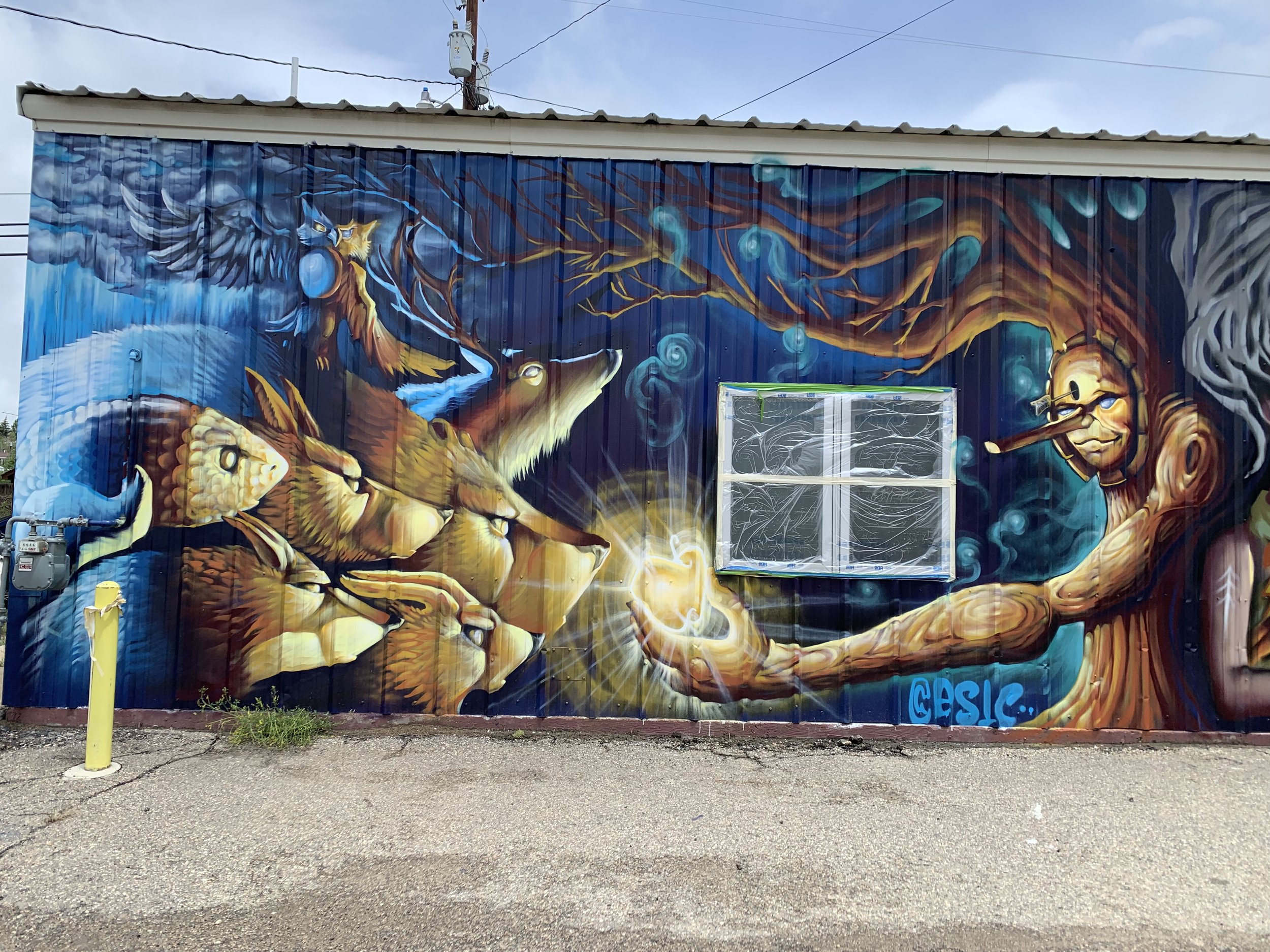 Colorful mural of a tree with a face holding a glowing orb in one hand. The tree's face has glasses and a pipe. The background is a starry night sky with clouds. There are animals, including a wolf and a fish, blending into the scene.