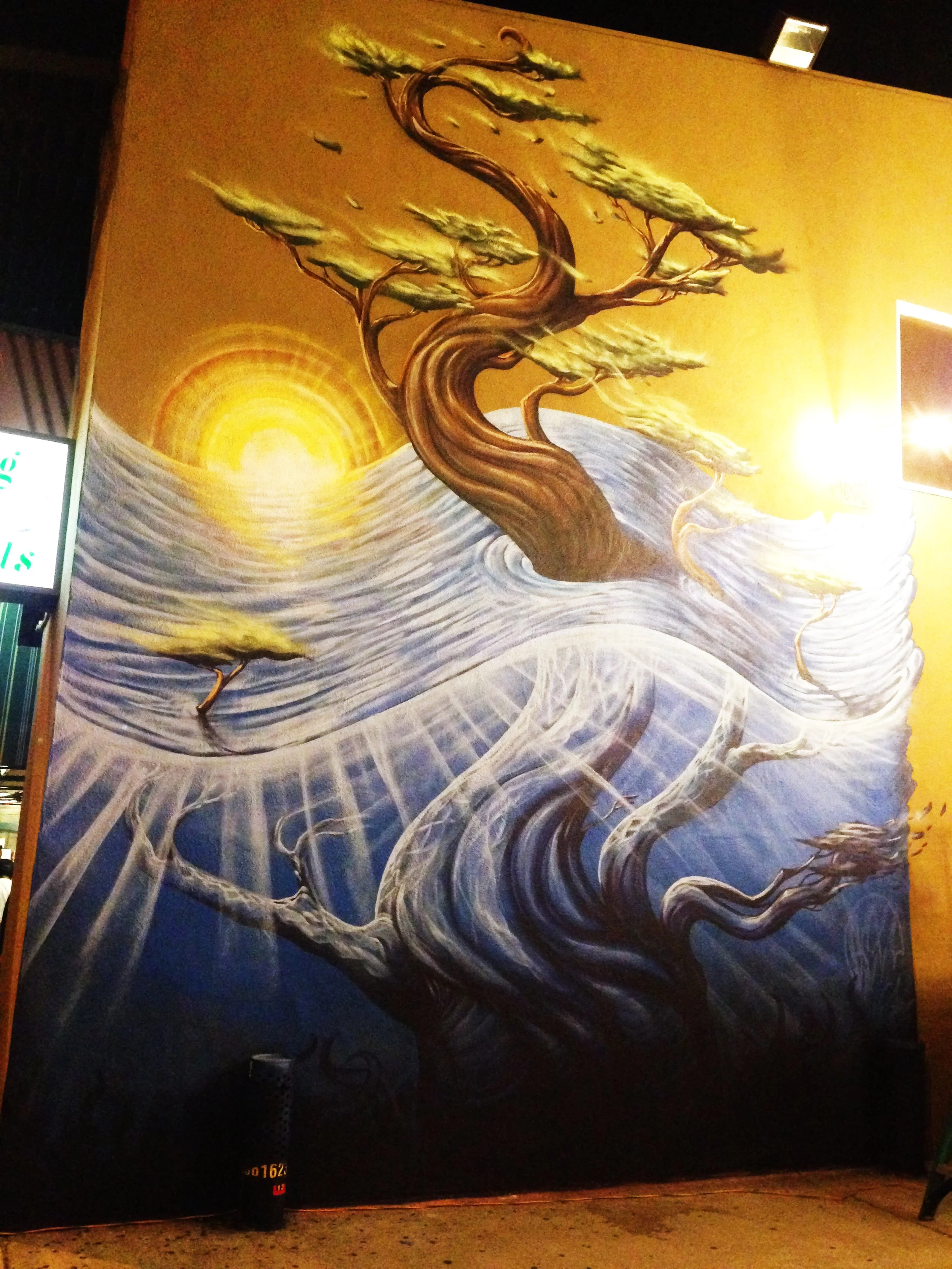 A large mural of a stylized tree with twisting branches and leaves, painted in tones of brown and green. The background features a bright yellow sun and sun rays, with a swirling sky and abstract waves.