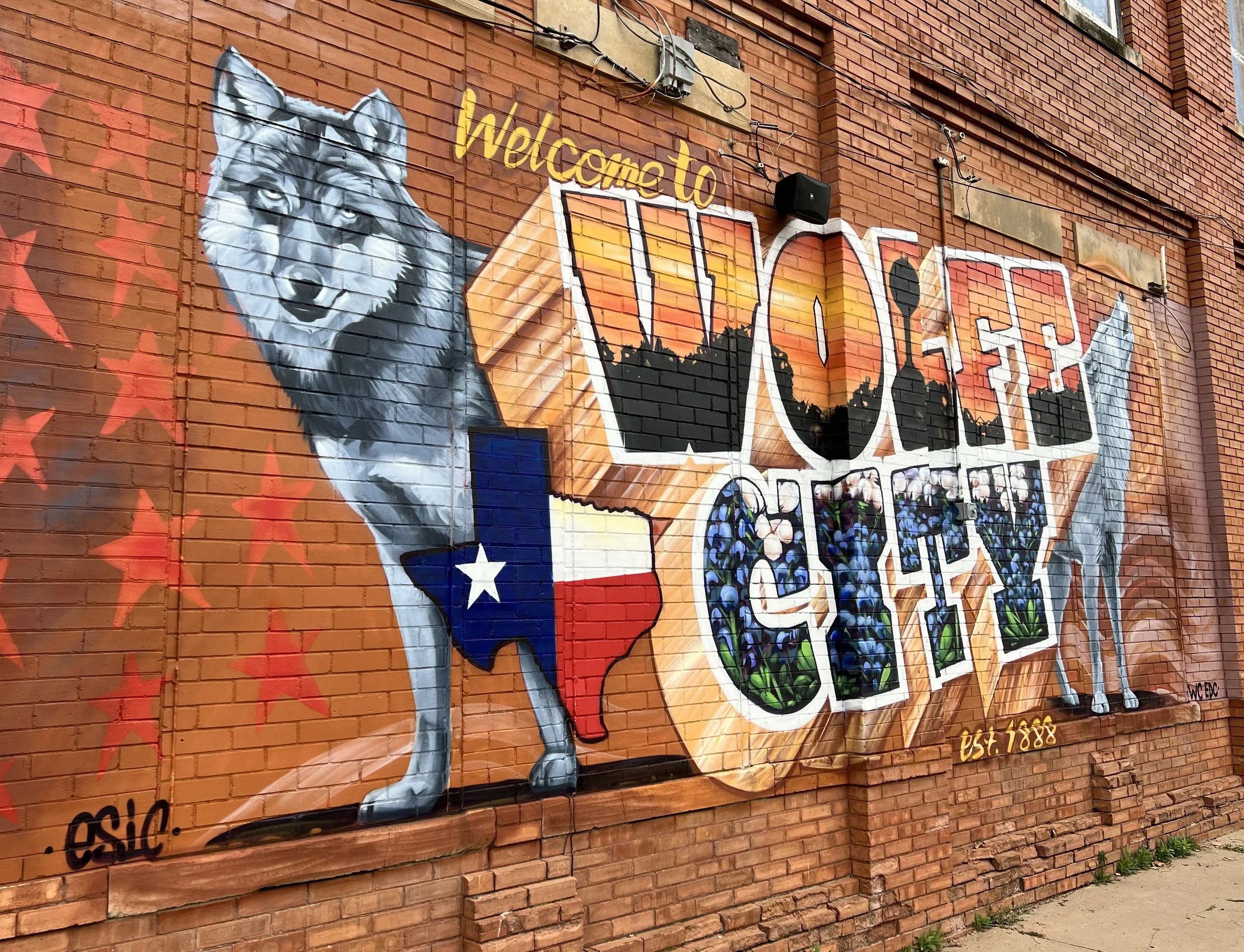 Colorful mural on a brick wall featuring a wolf and the Texas flag, with the words 'Welcome to Woes' and 'Est. 1888' written on it.