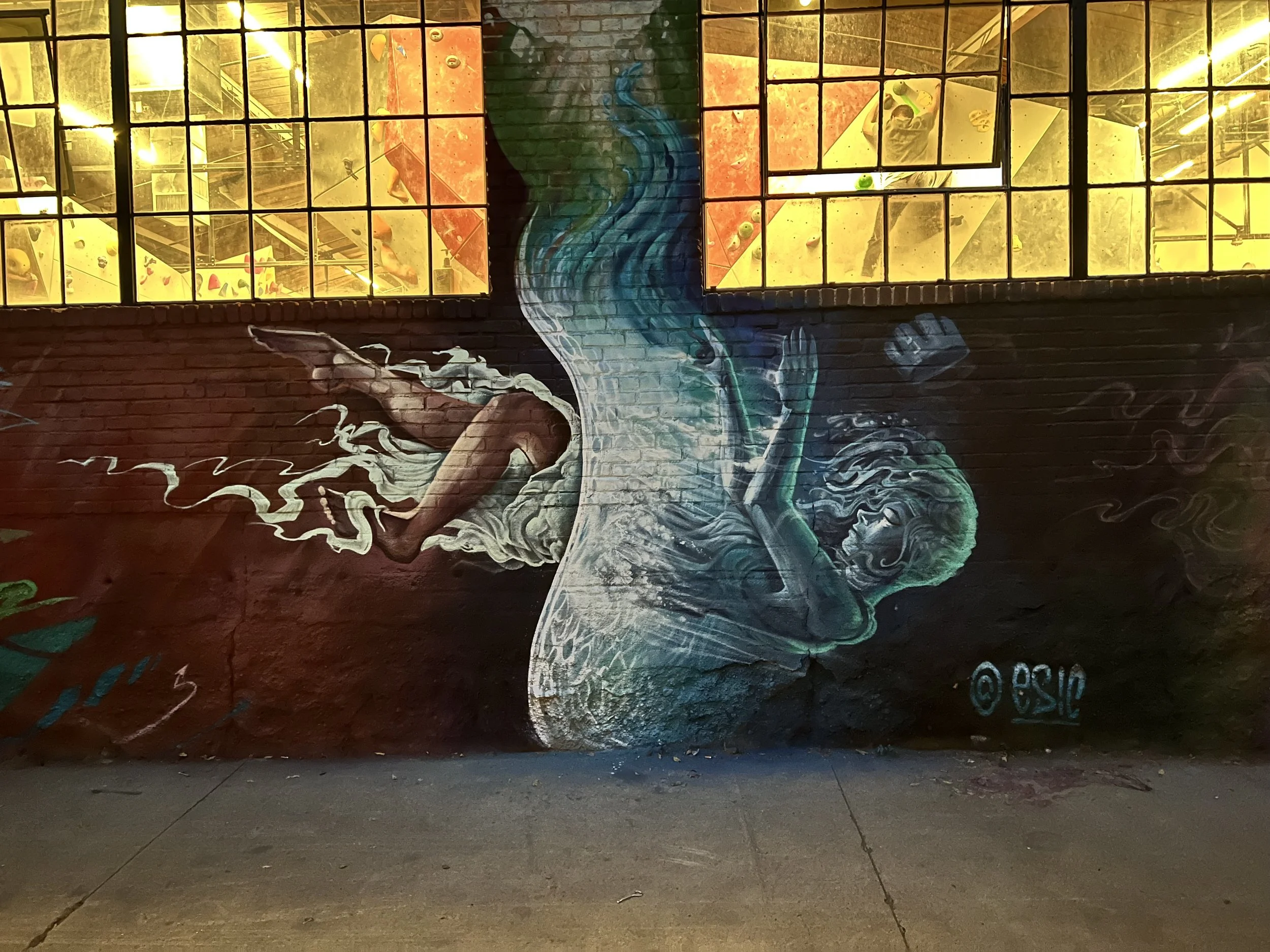 Street mural depicting a woman with blue hair, floating in water with her eyes closed, on a dark brick wall with illuminated windows above.