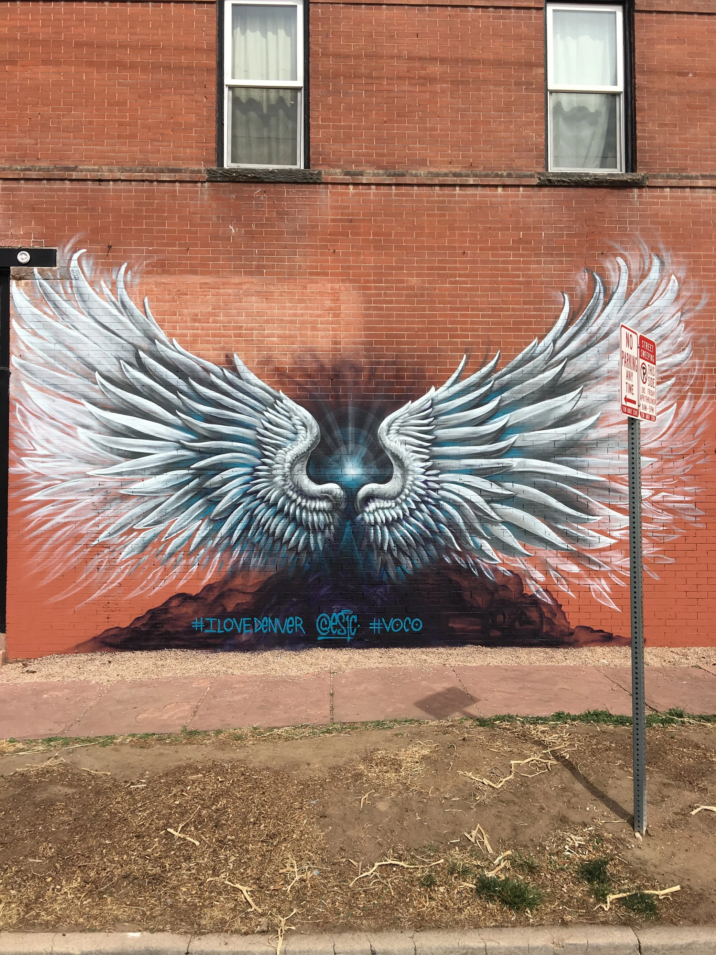 Street art mural of a bird with wings spread wide, painted on a brick wall. The wings are detailed with white, gray, and black feathers, and there are blue and purple hues at the base. Text on the mural reads #ILOVE DENVER @ESIC #VOCO.
