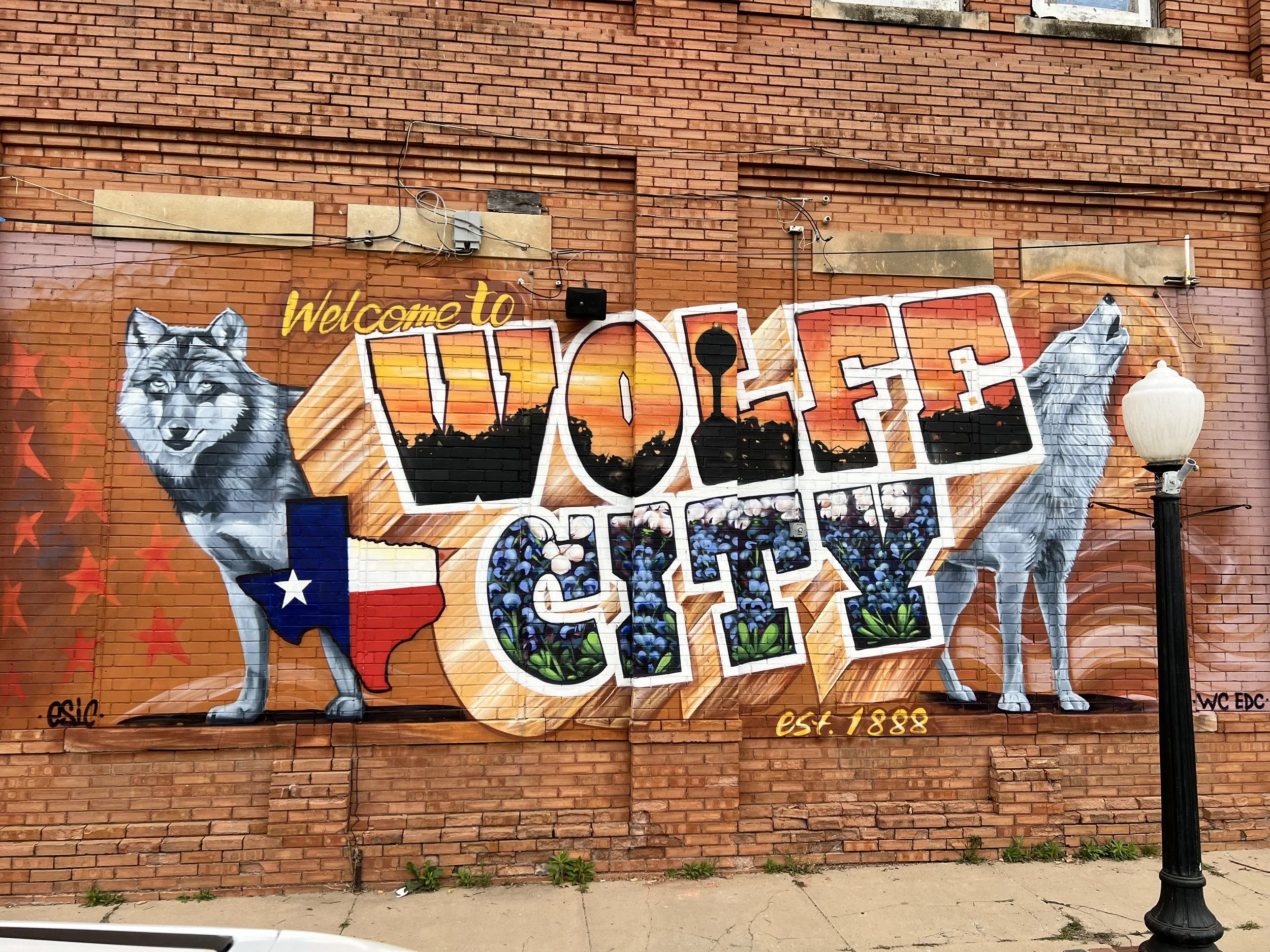 Colorful mural on brick wall featuring a wolf on each side, a Texas flag, and large stylized text reading "Welcome to Wolff City," with background elements including a sunset, city skyline, and flowers. A street lamp is visible on the right.