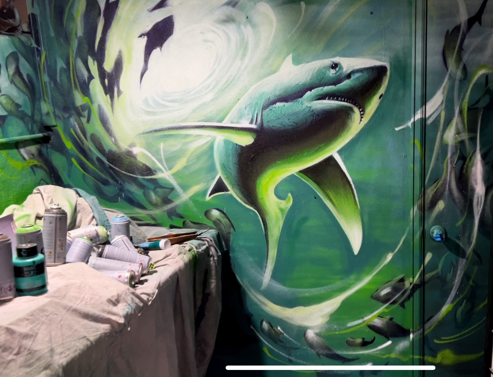 A colorful mural of a great white shark breaching water, painted on a wall. There are spray paint cans and art supplies on a cloth-covered table in front of the mural.