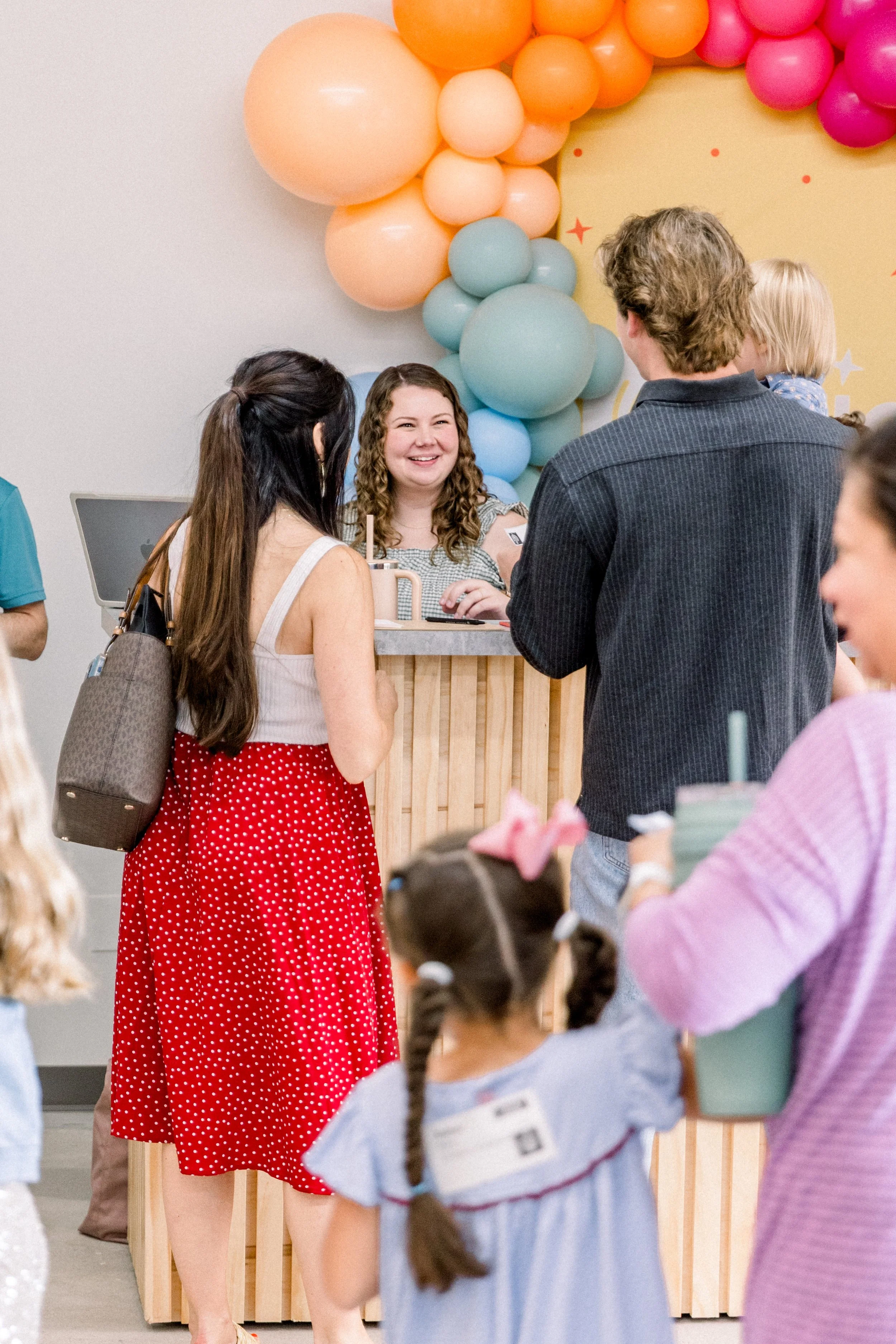 Group of people at a birthday party, with a woman in a red polka dot skirt talking to a woman behind a wooden counter decorated with pastel balloons.