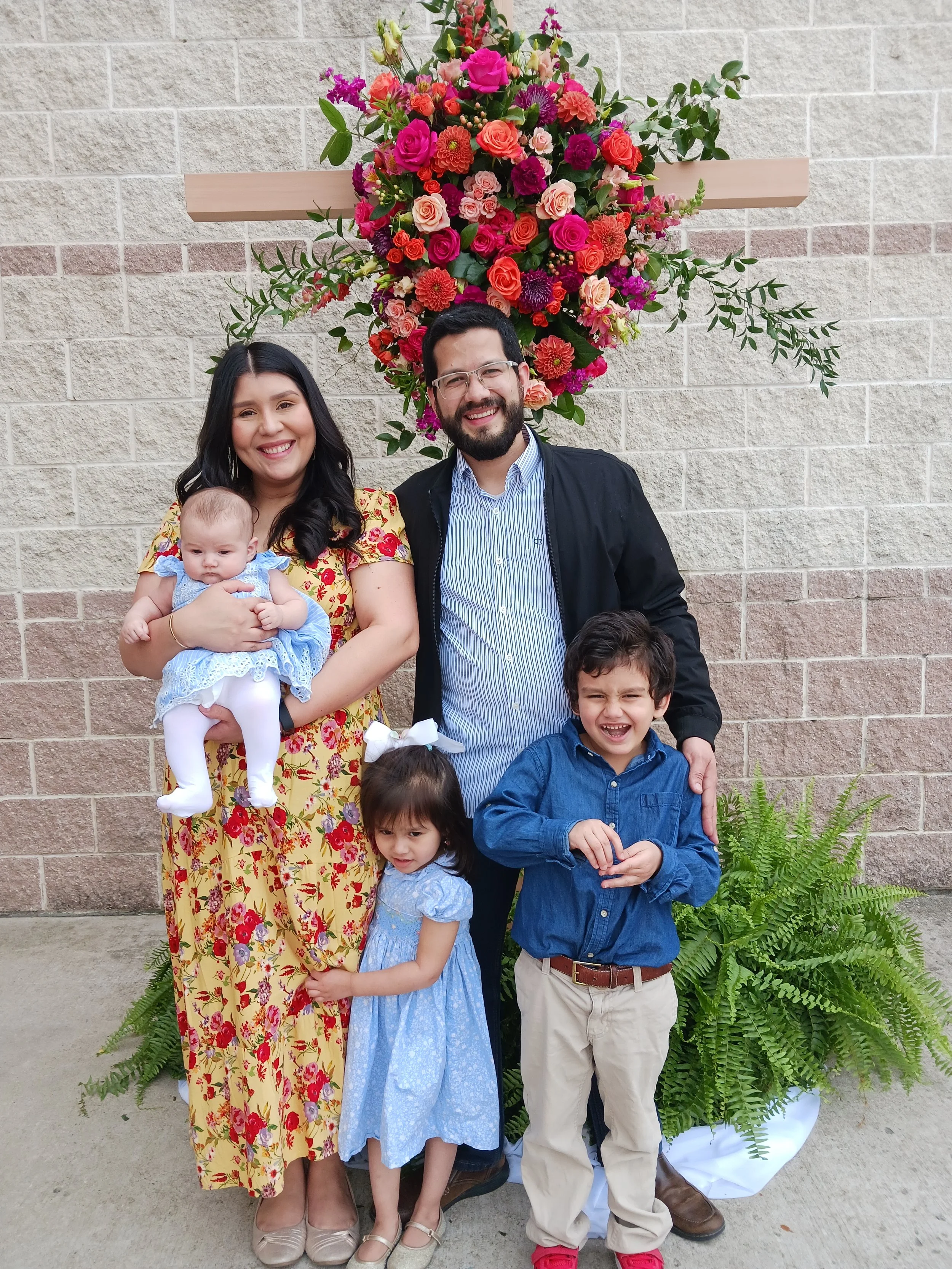 A family of five posing outdoors in front of a brick wall decorated with a large, colorful floral arrangement. The group includes two women, one holding a baby girl, and three young children, all smiling and dressed in colorful, casual attire.