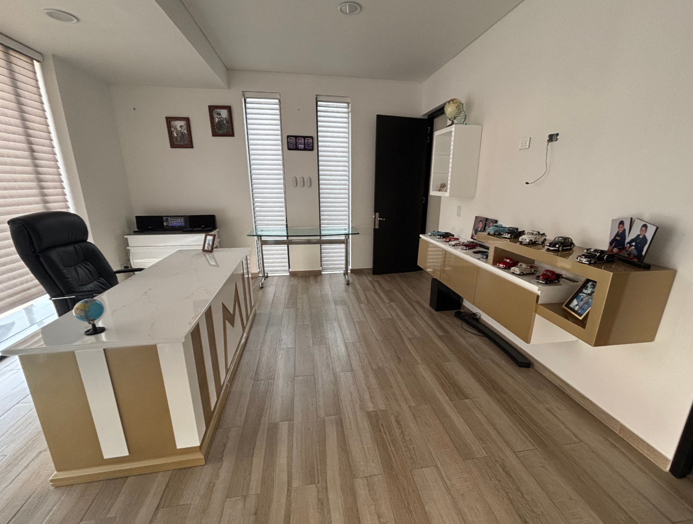 Despacho con escritorio blanco, silla negra, mueble con modelos de autos y fotos, ventanas con persianas, paredes blancas con cuadros y armario
