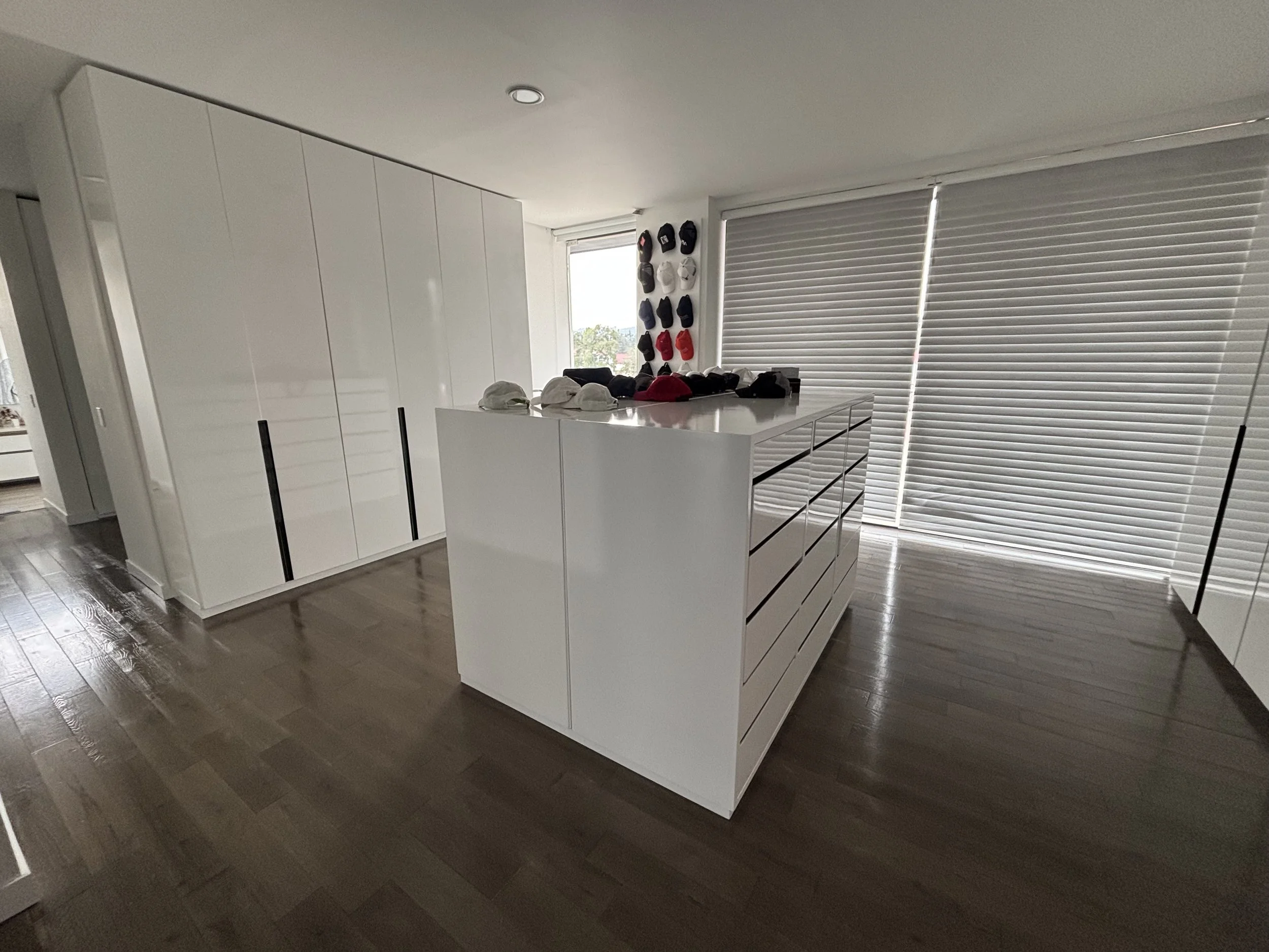Cocina moderna con gabinetes blancos, ventana grande con persianas cerradas, suelo de madera oscura y un mostrador central con ropa y accesorios en la superficie.
