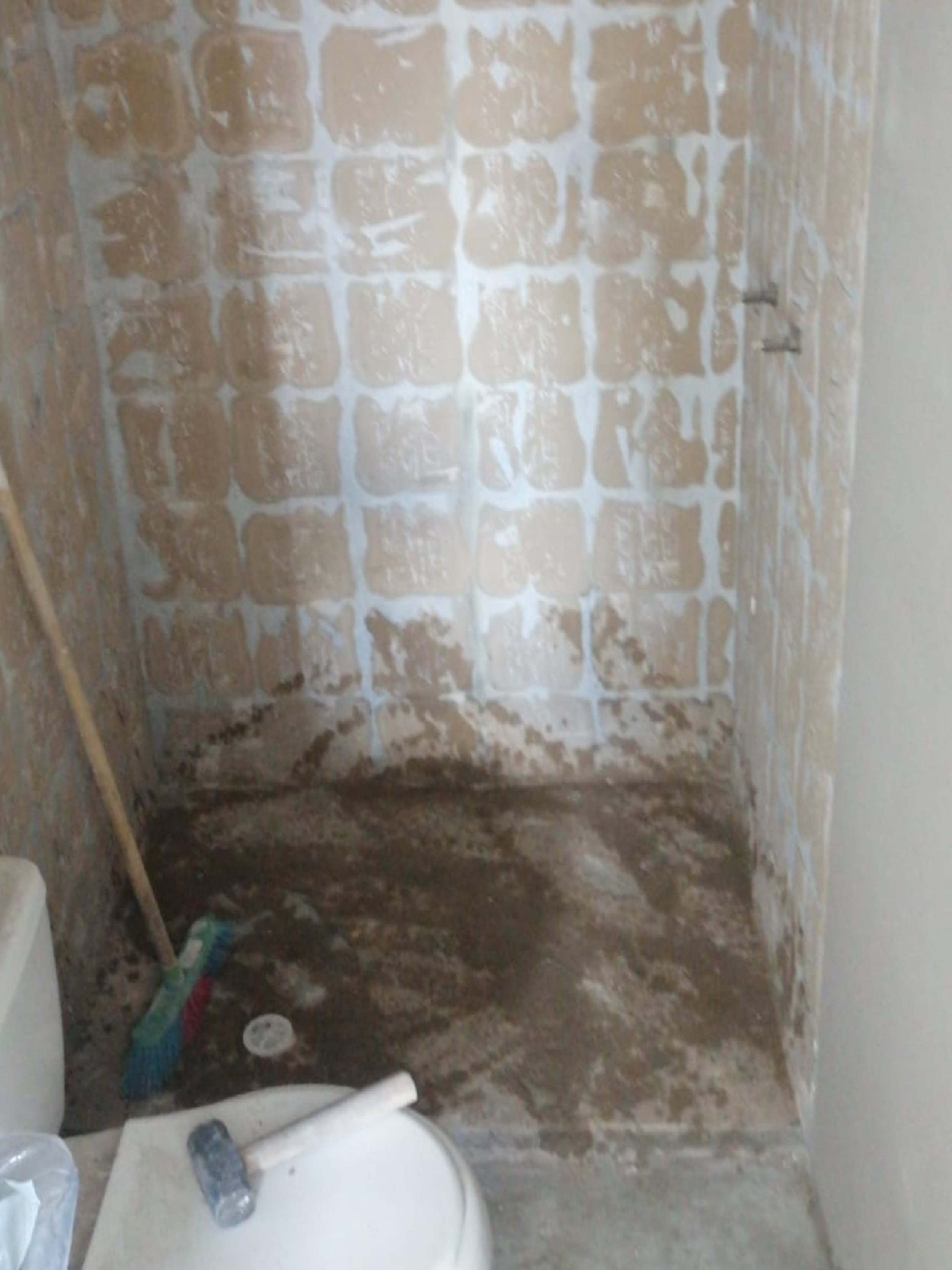 Un baño en construcción con paredes de bloques de cemento, suelo de tierra húmeda y algunas herramientas como escoba y mascarilla en el suelo.