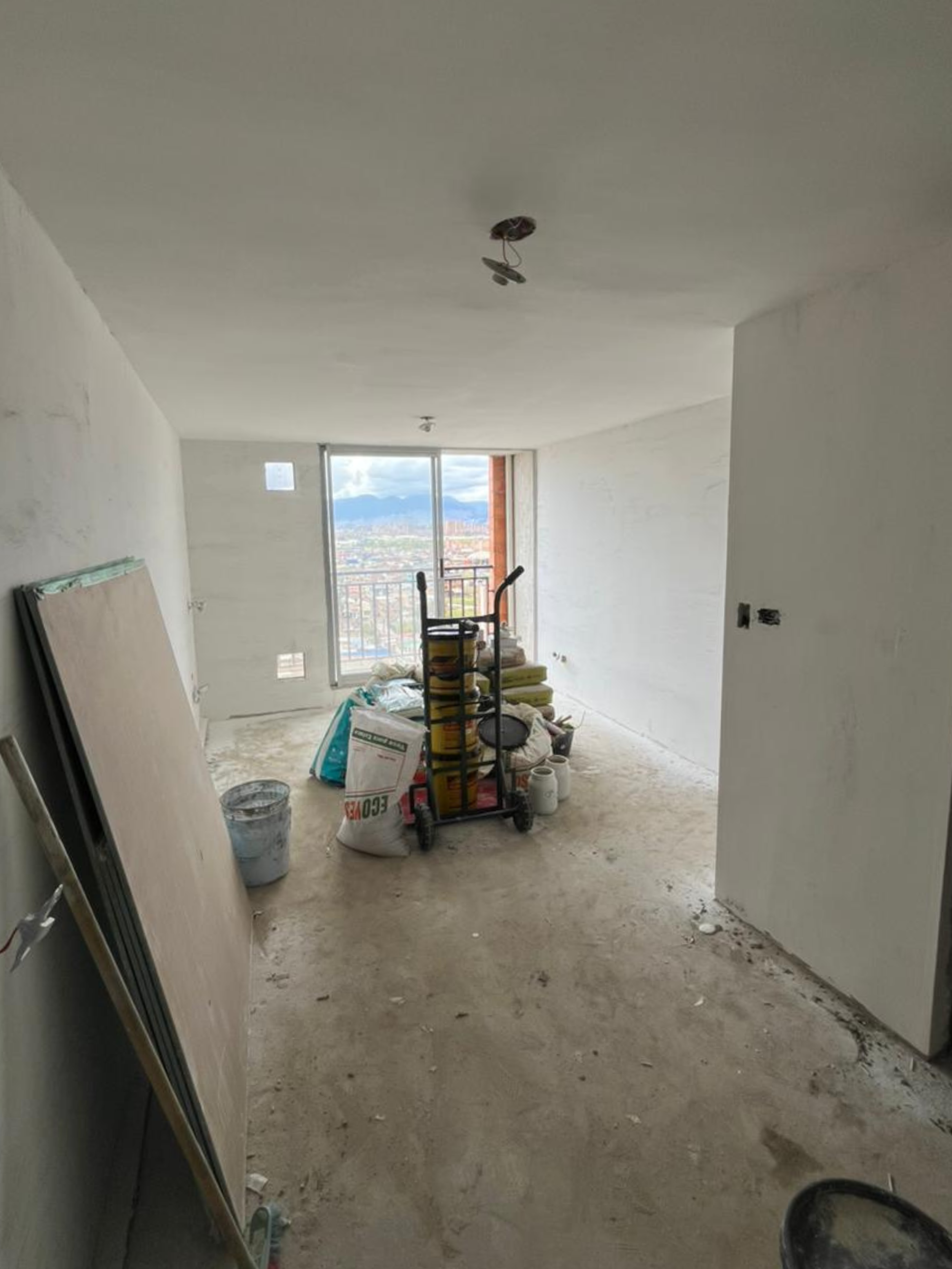 Interior de un apartamento en construcción con paredes blancas, piso de concreto, y una ventana grande que da vista a la ciudad y a montañas lejanas. En el centro hay una carretilla con materiales y herramientas de construcción, y en el suelo hay bol
