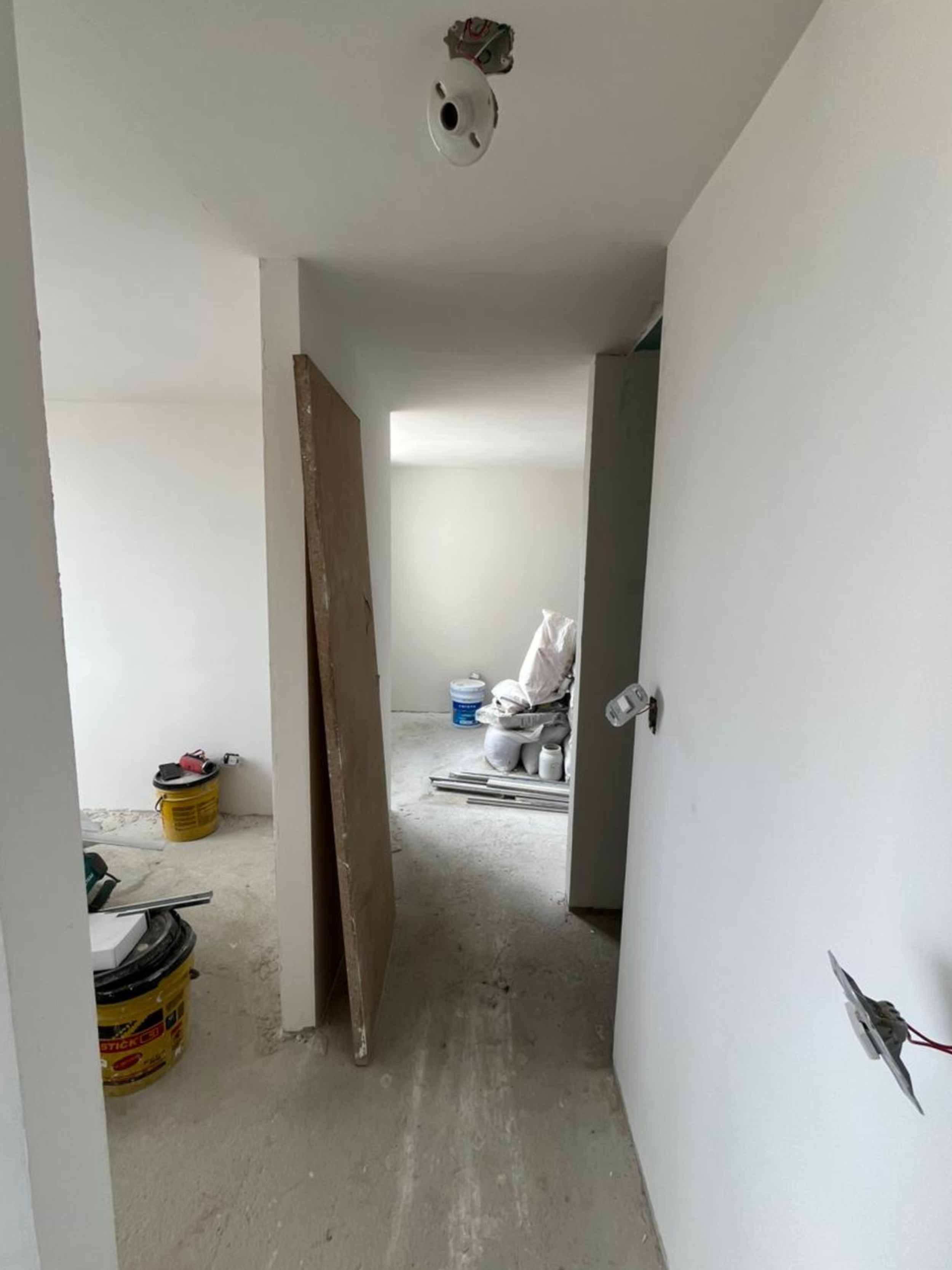 Interior en construcción con paredes blancas, piso sin acabado y materiales de construcción apilados, incluyendo cubetas de pintura, madera y cables eléctricos.