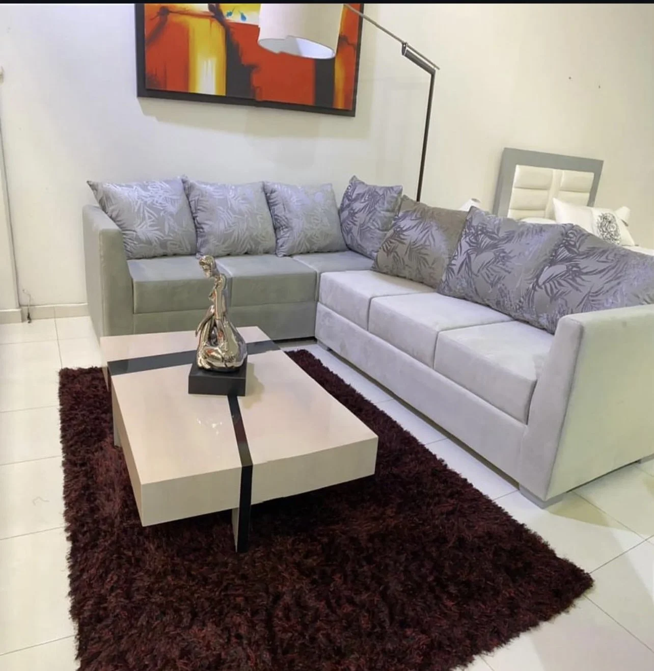 Sofá gris con cojines decorativos en una sala, mesa de centro con una escultura metálica y alfombra roja. Pintura abstracta en la pared y lámpara de pie.