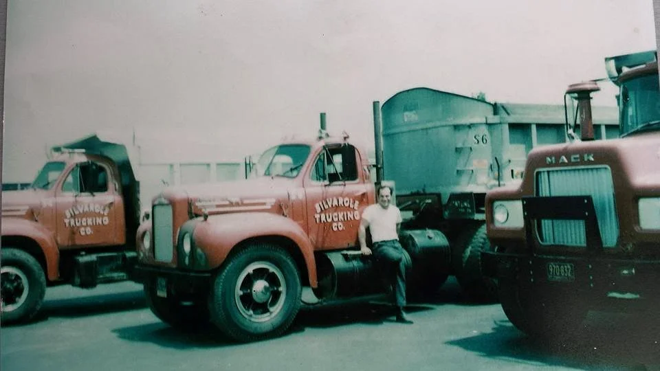 A person leaning against a vintage truck with a cargo container, parked between two other trucks in a lot.
