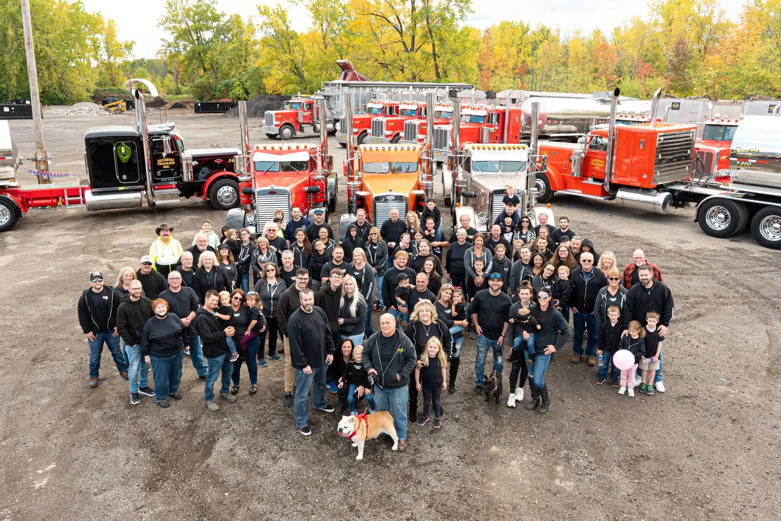 Group photo of people gathered in front of vintage fire trucks outdoors during autumn, with colorful trees in the background.